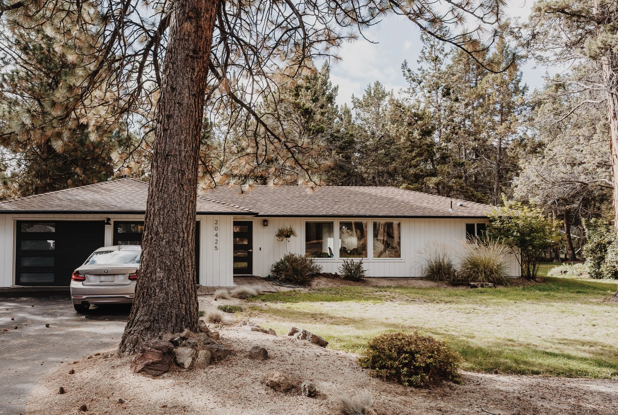 Front view of a house with large windows, surrounded by tall pine trees, with a car parked in the driveway and a small garden in front.