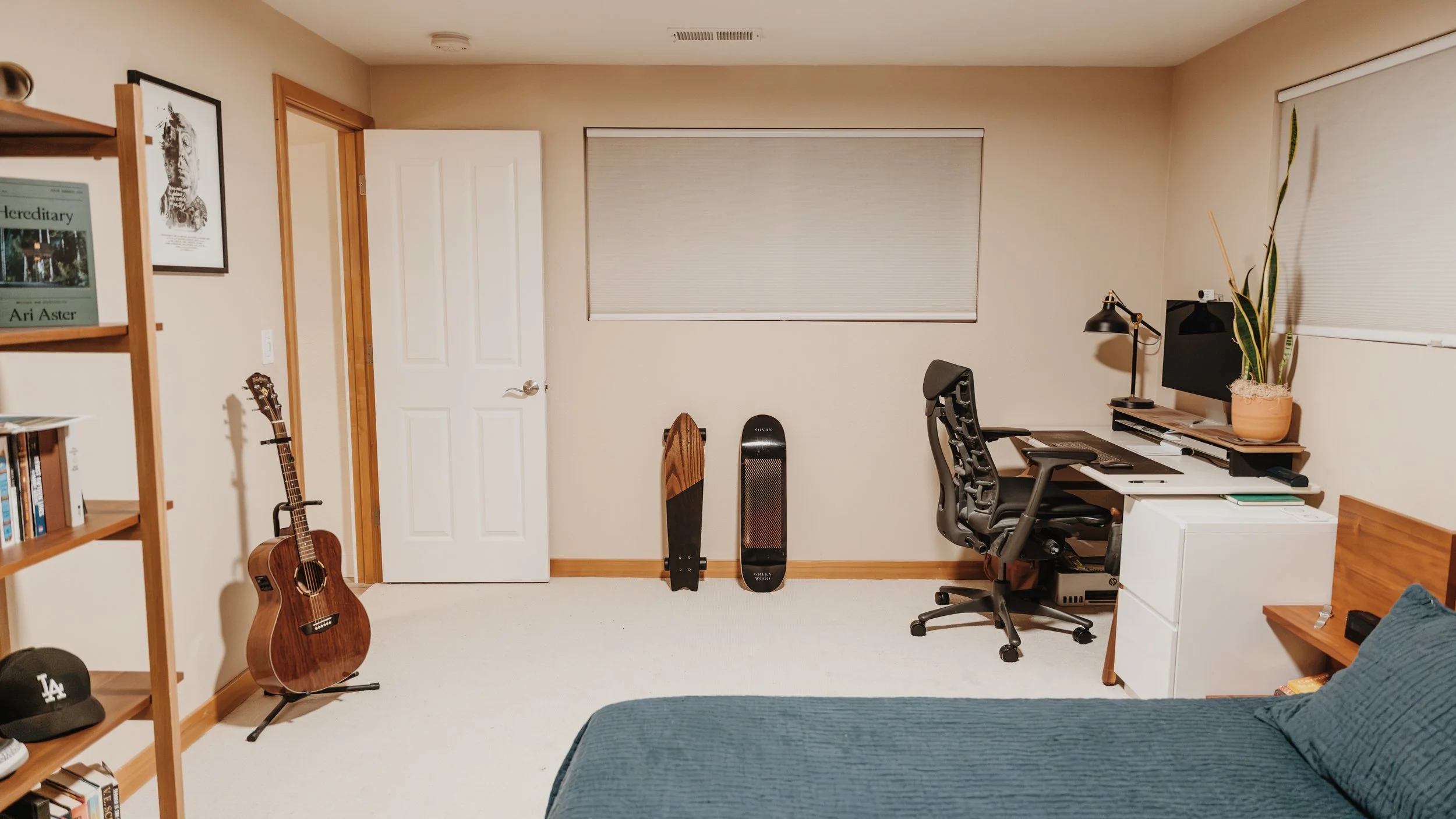 A cozy home office with a wooden desk, black ergonomic chair, computer monitor, plants, skateboard, acoustic guitar, and bookshelves.