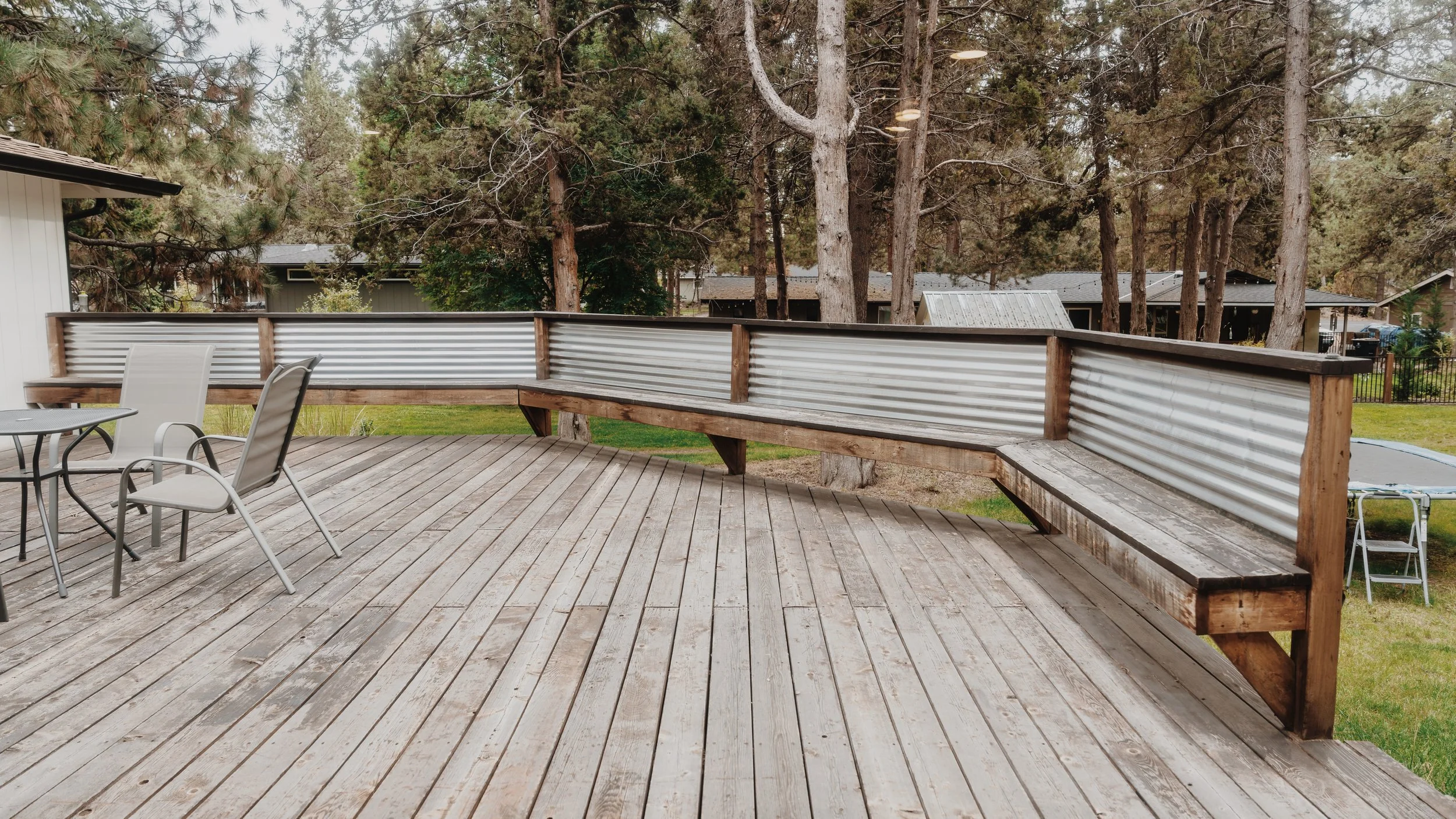 A wooden porch with metal railing and outdoor seating in a backyard with trees and neighboring houses.