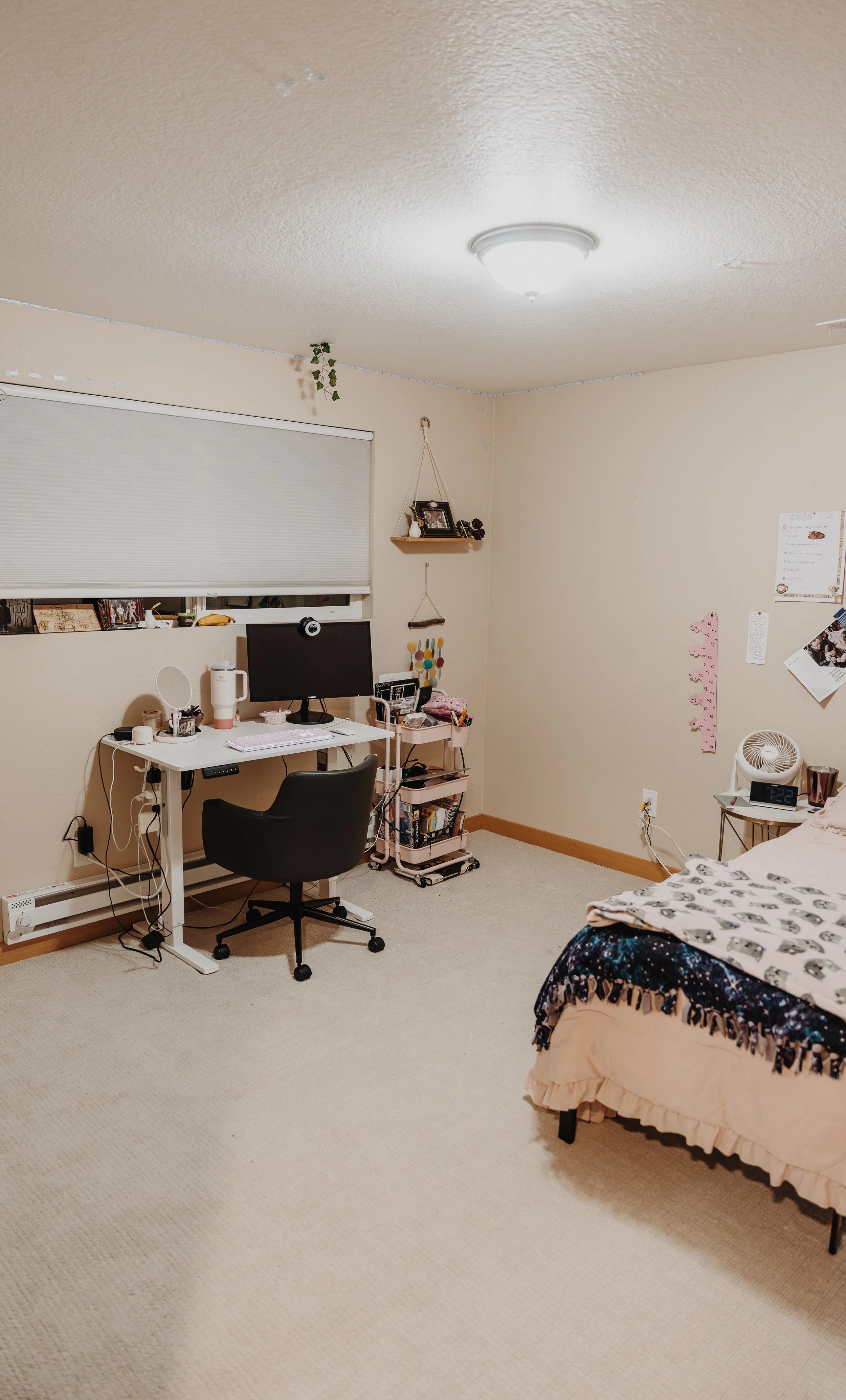 A bedroom with beige carpet, cream-colored walls, a white desk with a black office chair, a computer monitor, and various items on the desk. There is a window with a closed blind, a wall shelf with framed photos and decorations, and a pink rolling cart with books. The bed has a patterned blanket, and there are pictures and notes on the wall. An alarm clock on a small table shows 1:22.