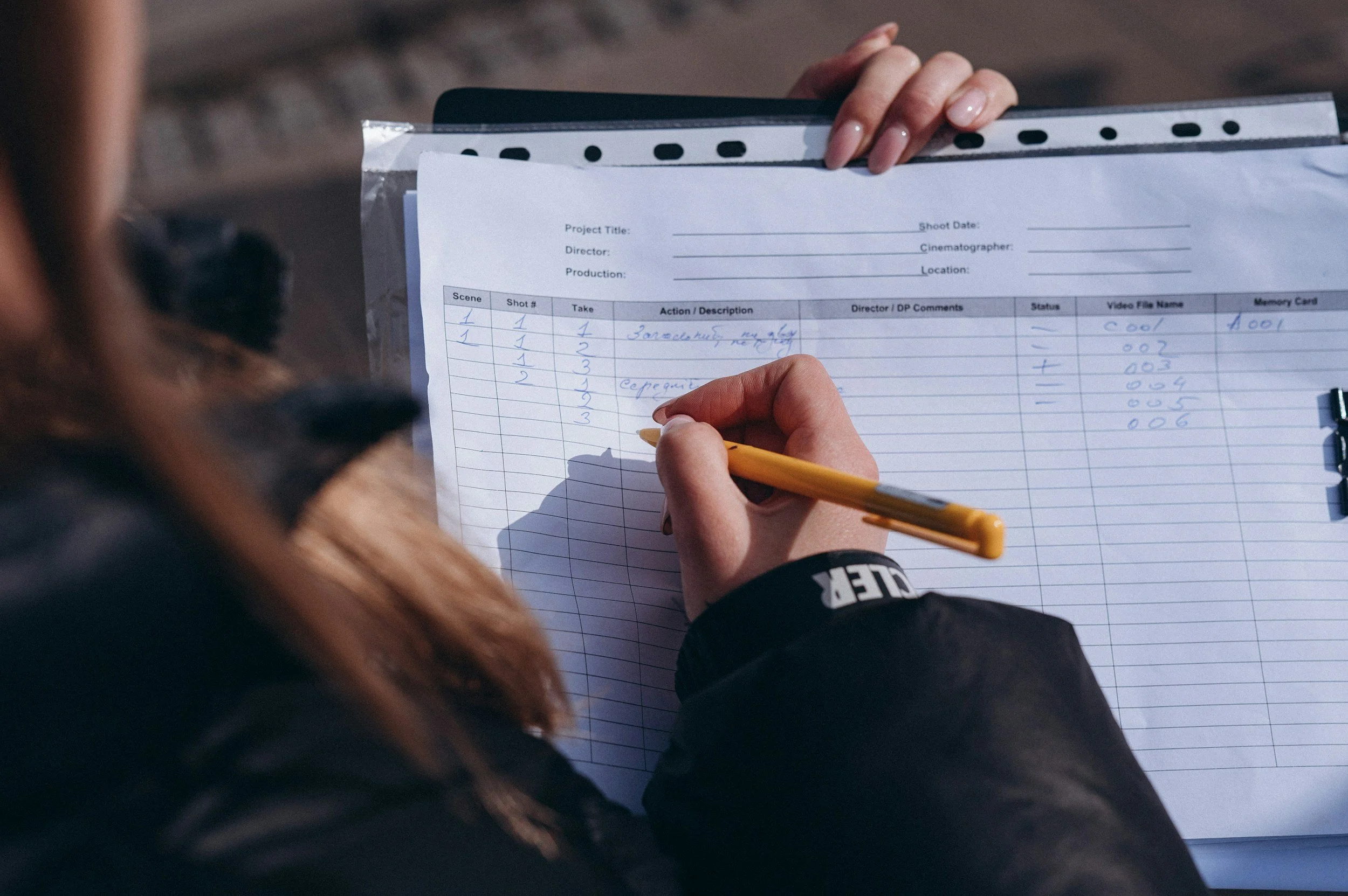 Close-up of a person's hand holding a yellow pen and writing on a production script or storyboard sheet attached to a clipboard, with a second person blurred in the background.