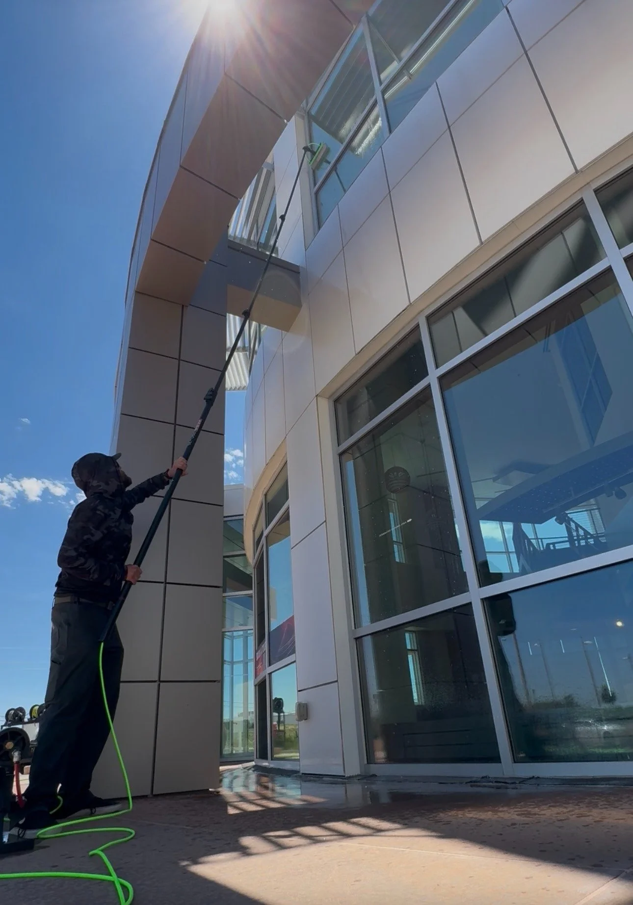 A person in dark clothing is cleaning a tall building's glass windows using a long-handled squeegee on a sunny day.