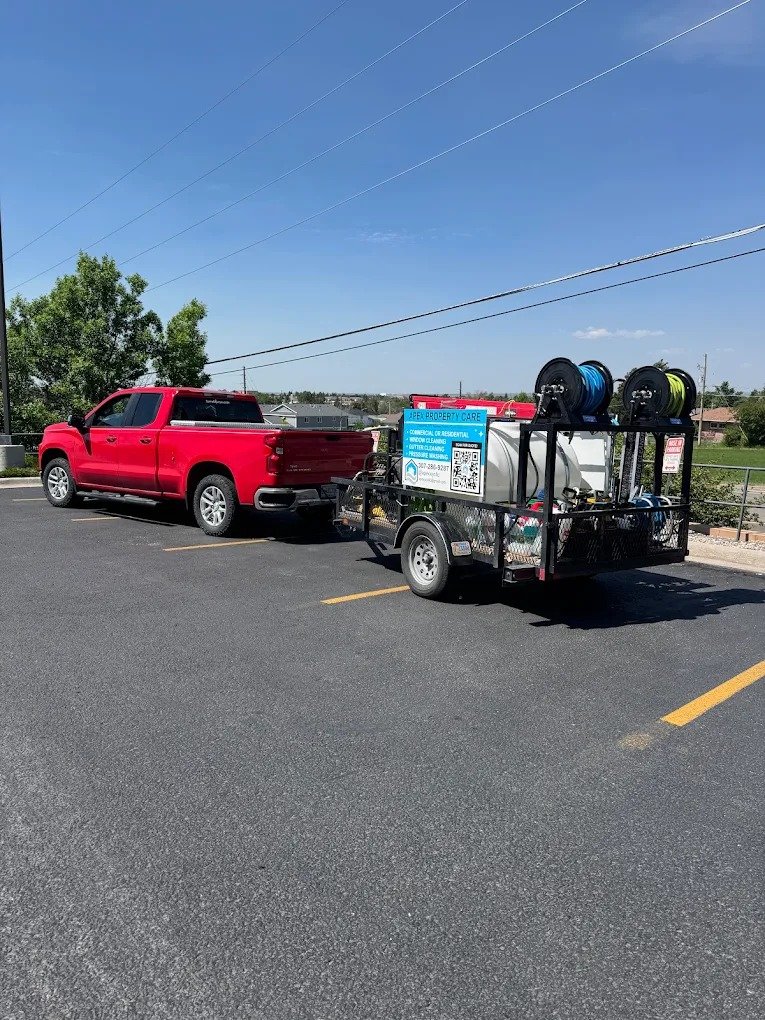 Red pickup truck towing a property care trailer with hoses and a sign, parked in an outdoor lot under a clear blue sky.
