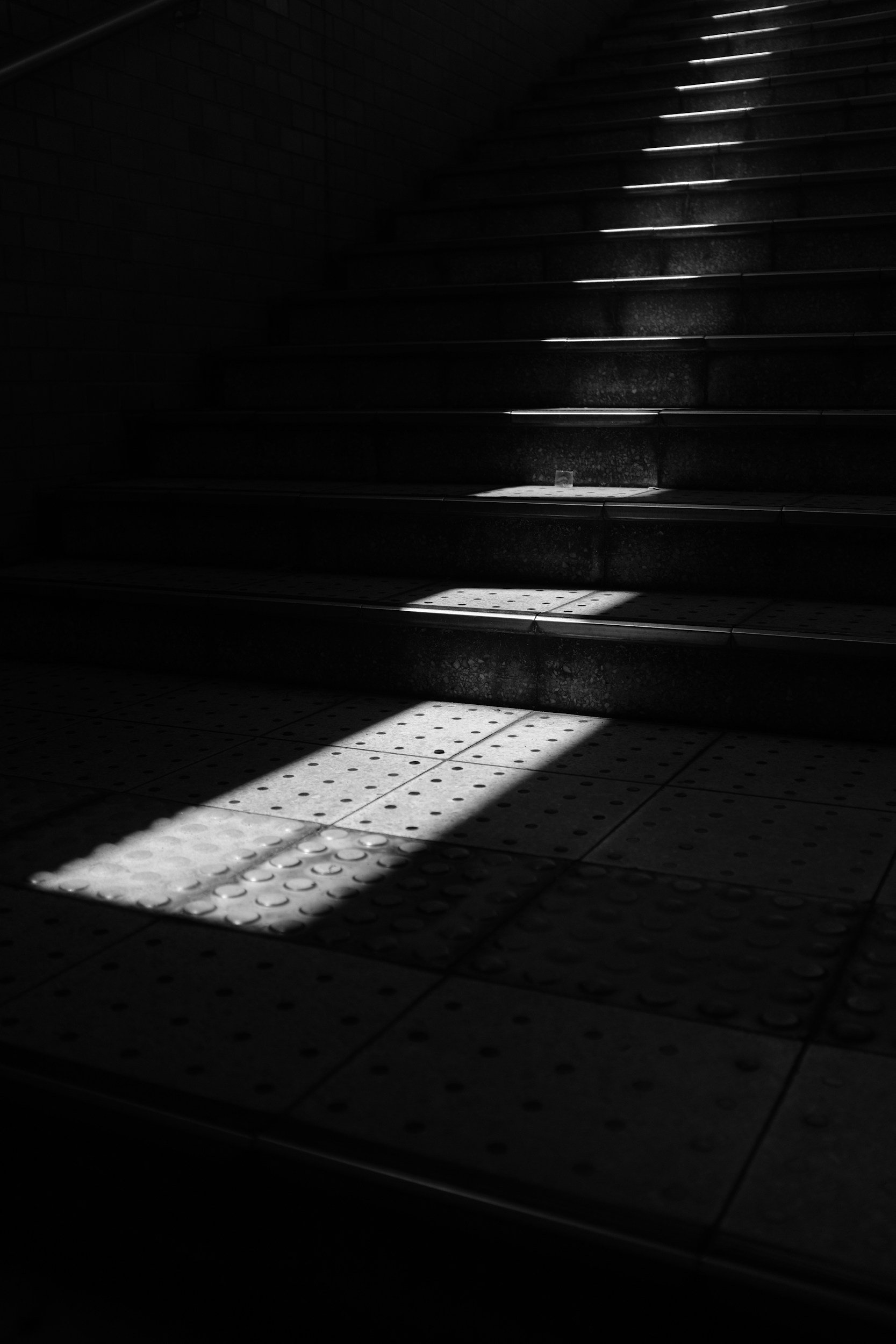 A staircase with dark steps and a brick wall on the left. Light filters in from the right, casting shadows on the steps and the tiled floor.