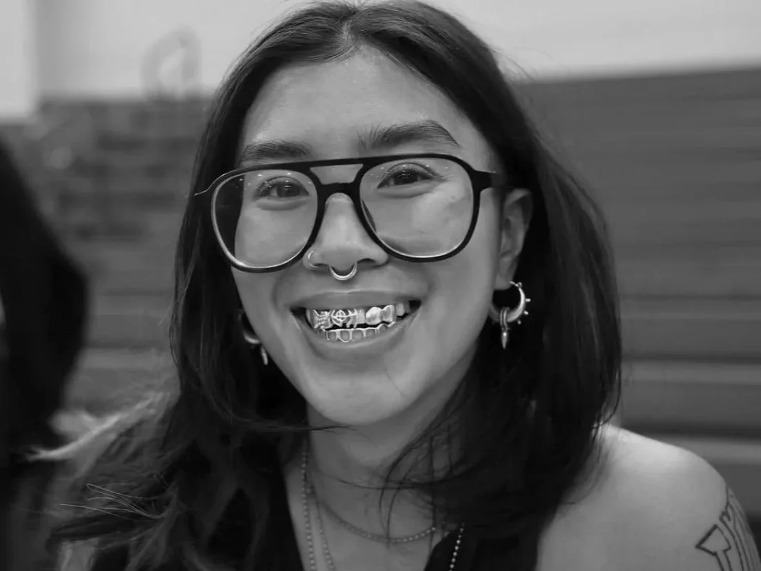 Close-up of a woman with shoulder-length dark hair, wearing glasses with large round frames, multiple earrings, and a septum piercing, smiling in black and white.