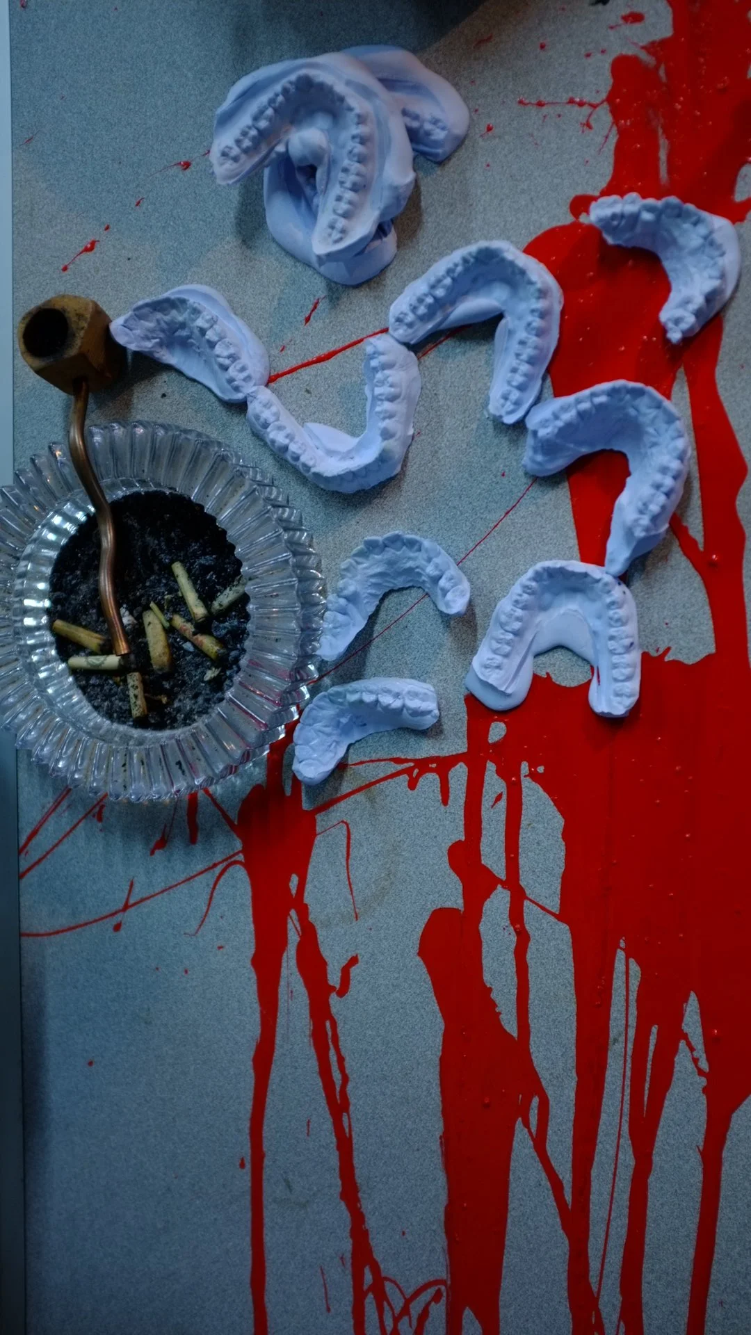 Casual scene with dental moldings arranged on a table, red liquid resembling blood splattered across the surface, and an ashtray with burnt cigarettes.