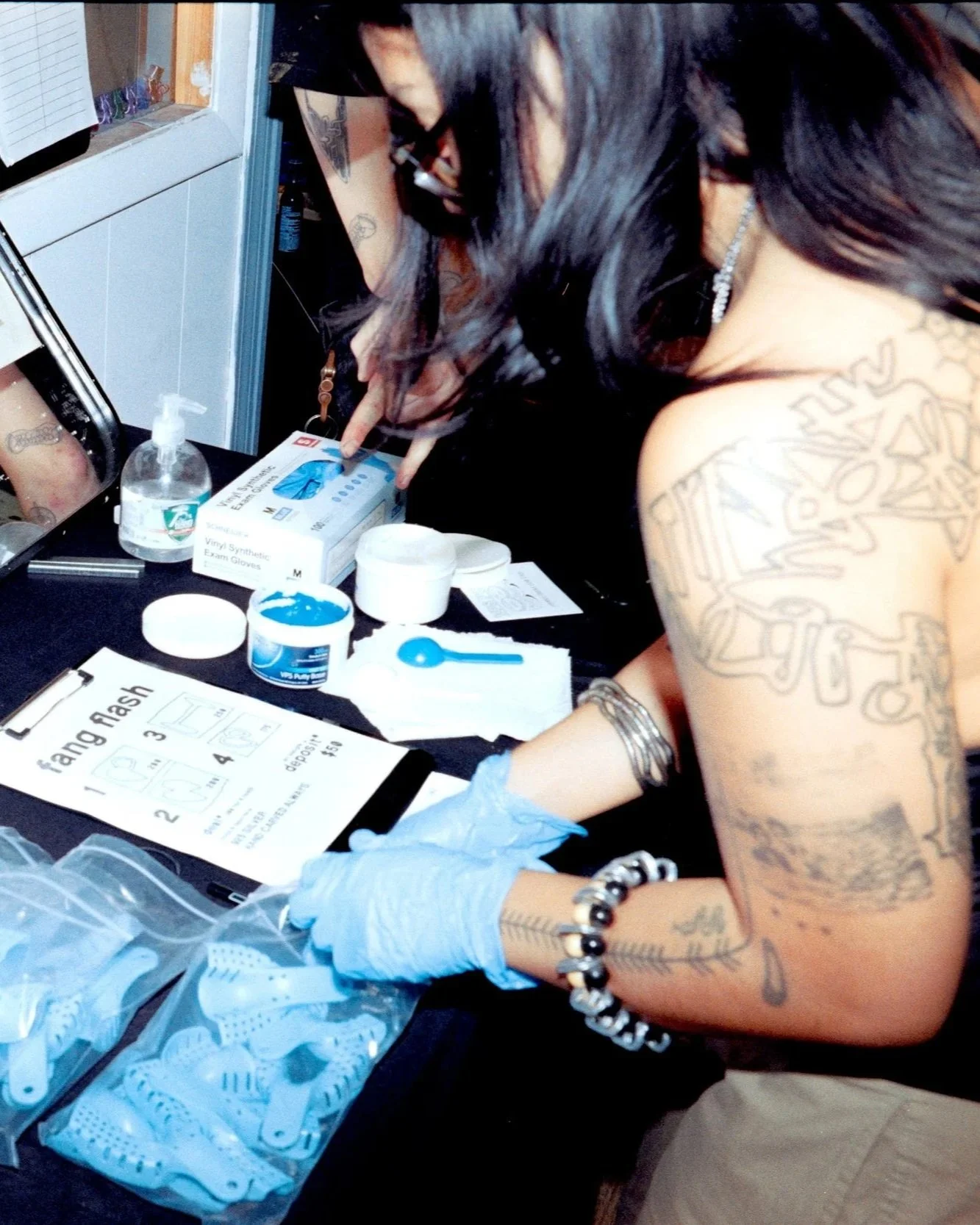 Person with tattoos and jewelry preparing medical or tattooing supplies, including latex gloves, vinyl exam gloves, and tattoo transfer papers on a black table.