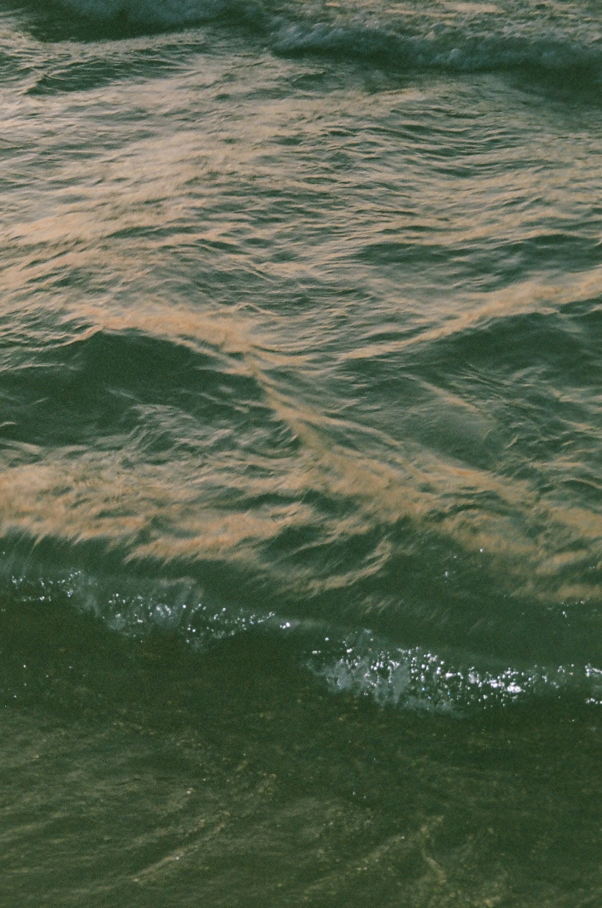 Close-up view of Lake Michigan with gentle waves and ripples in shades of dark green and tan, with small splashes at the bottom.