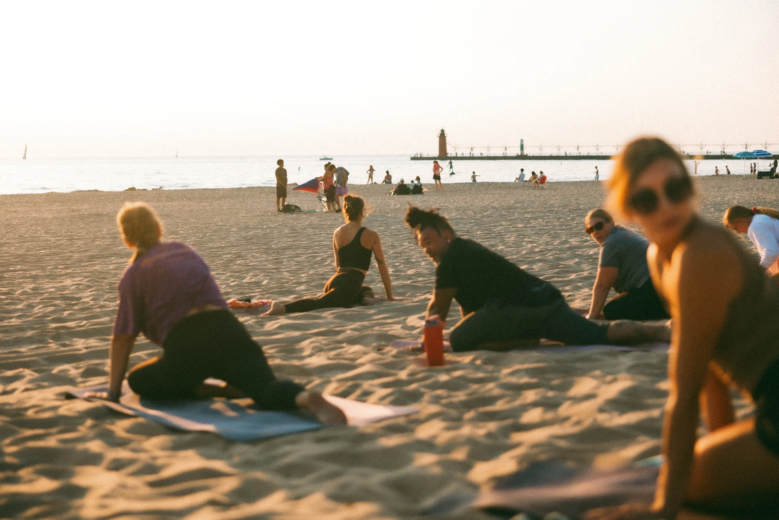People relaxing and exercising on a sandy beach during sunset, with a lighthouse and Lake Michigan in the background.