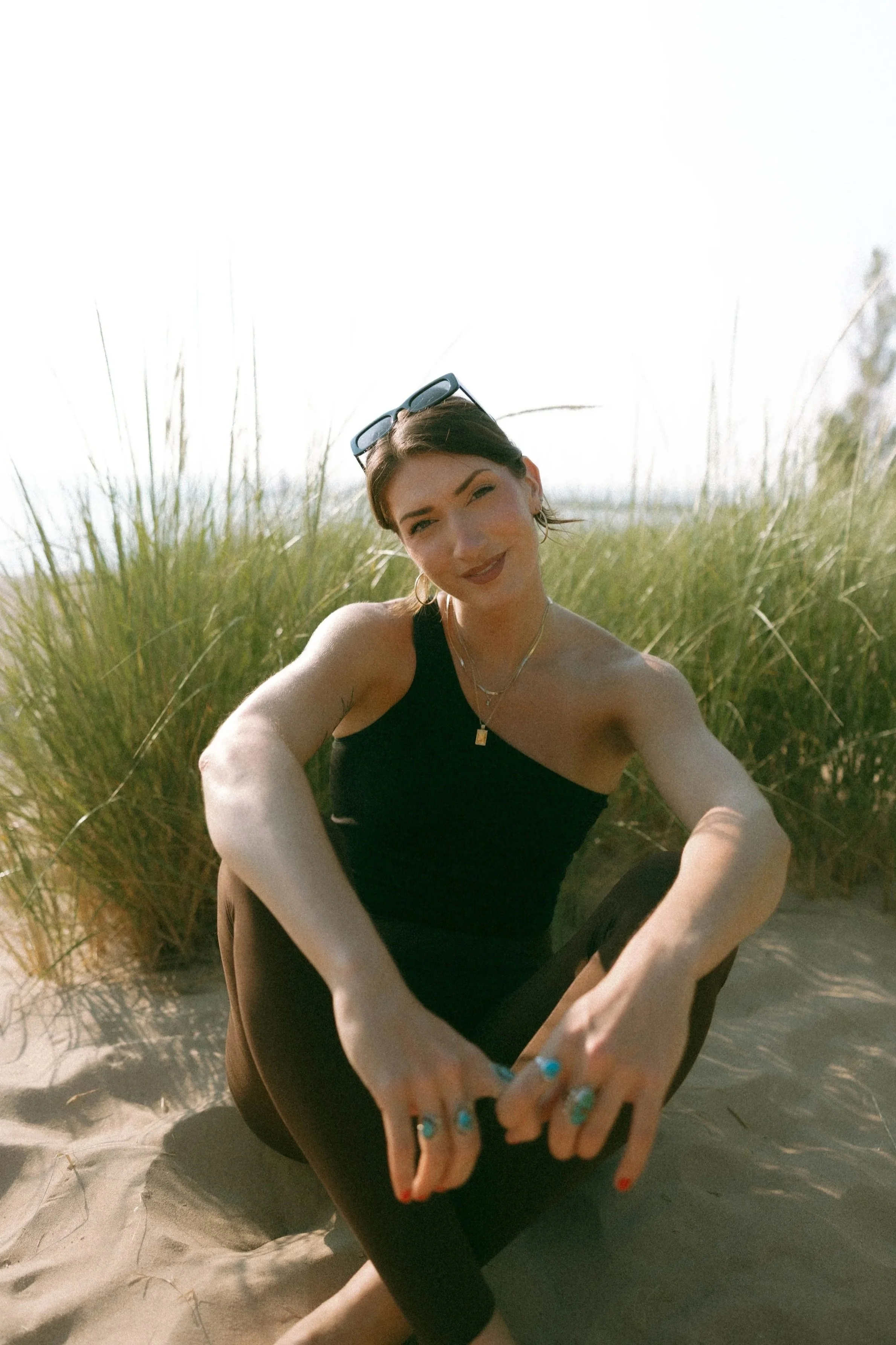 A woman sitting in sand surrounded by tall grass, wearing a black tank top and sunglasses on her head, smiling.
