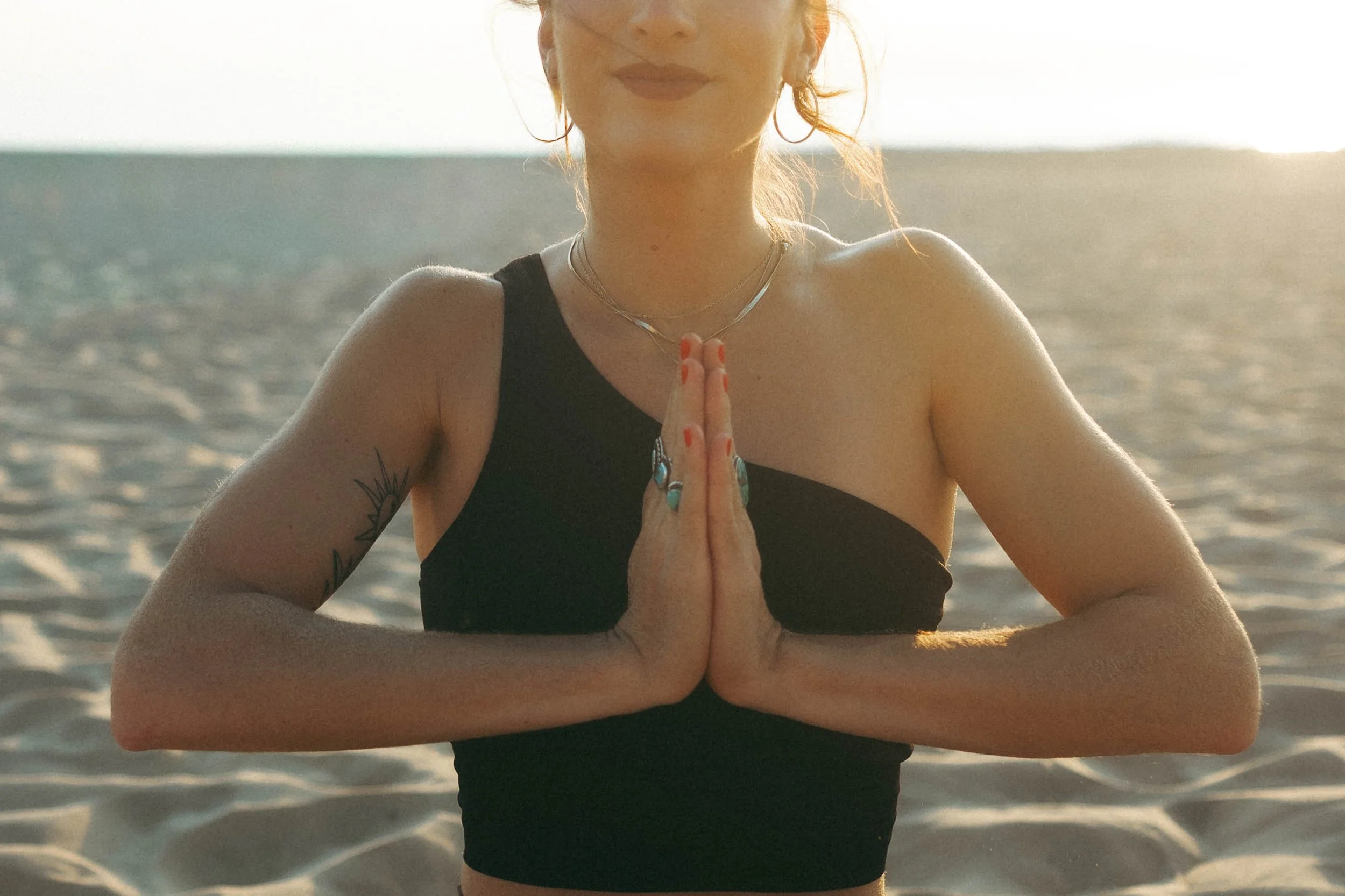 A woman practicing yoga on a sandy beach during sunset, with her hands in a prayer position at her chest.