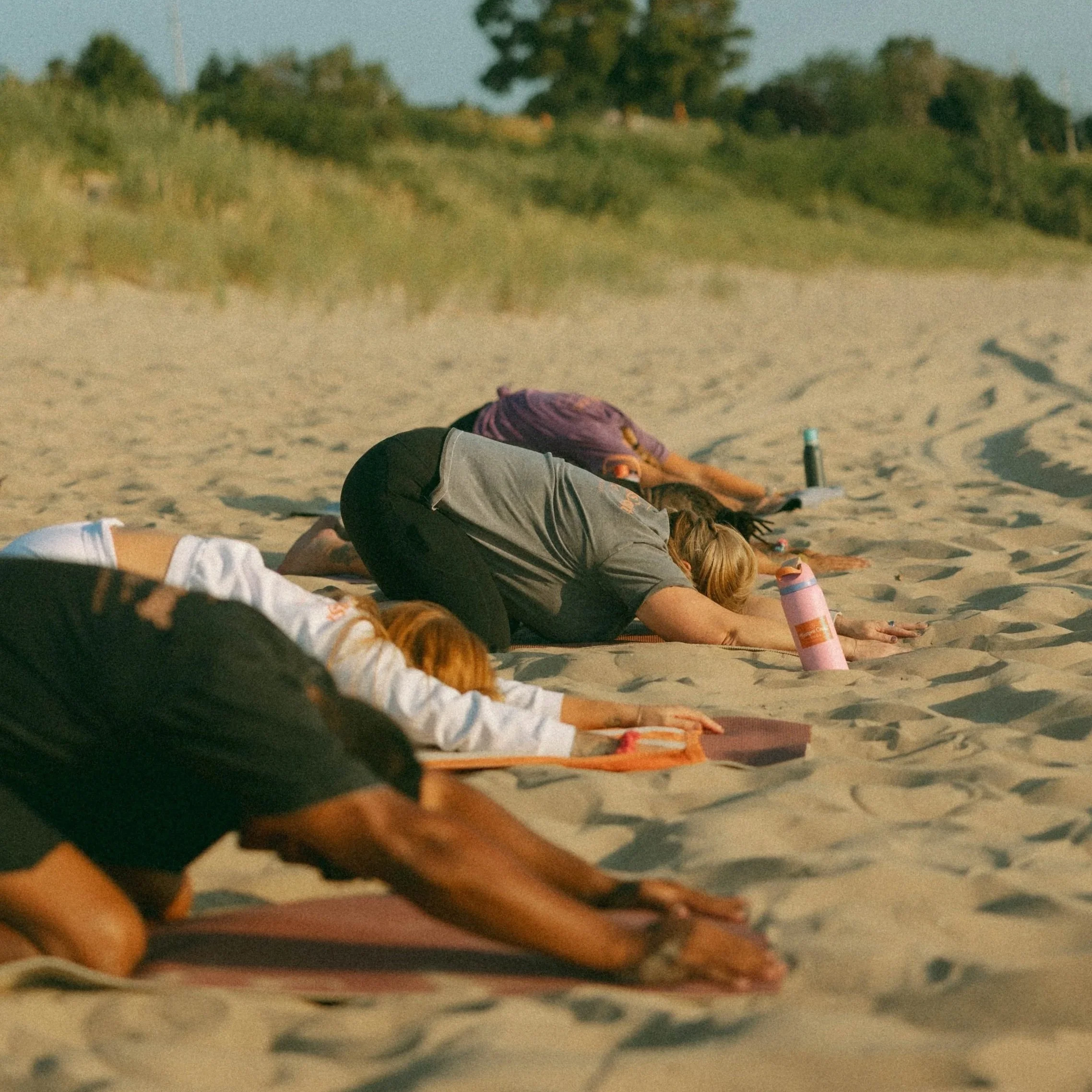 People practicing yoga on a sandy beach, lying on mats and in poses, with trees and a hill in the background.
