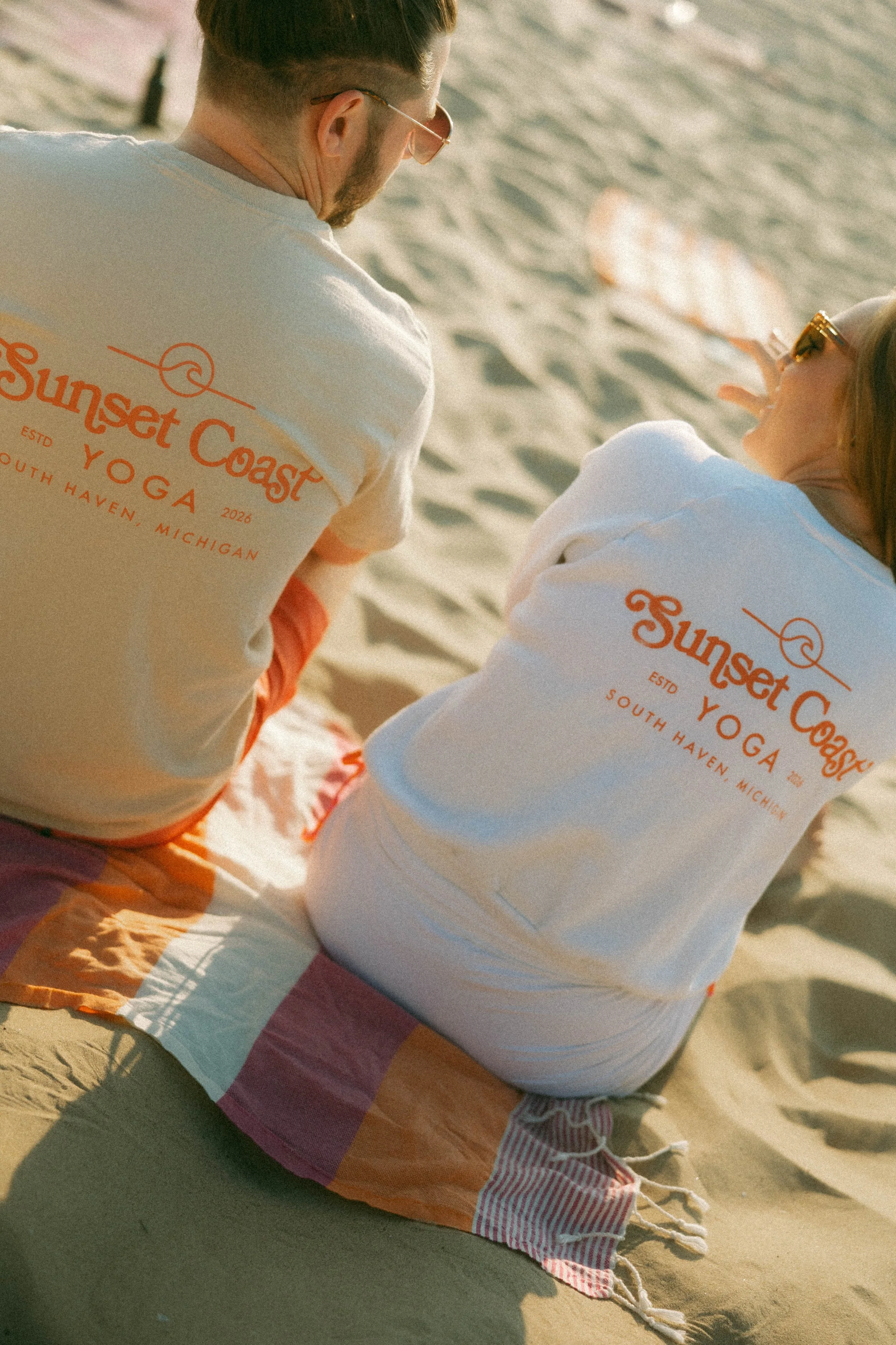 A man and woman sitting on a sandy beach during sunset, wearing matching white t-shirts with orange text that reads 'Sunset Coast Yoga South Haven, Michigan'. The man has tattoos on his arm and is wearing glasses, while the woman has sunglasses and is sitting with her back turned to the camera.
