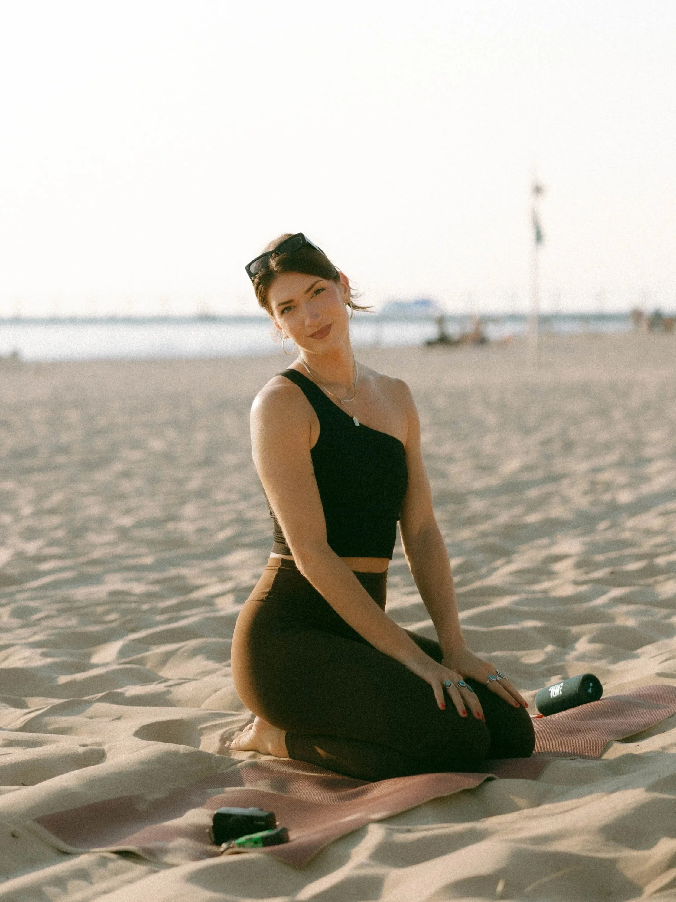 A woman with short hair and sunglasses on her head is sitting on a yoga mat on the beach, smiling at the camera, with the ocean and sandy shoreline in the background.
