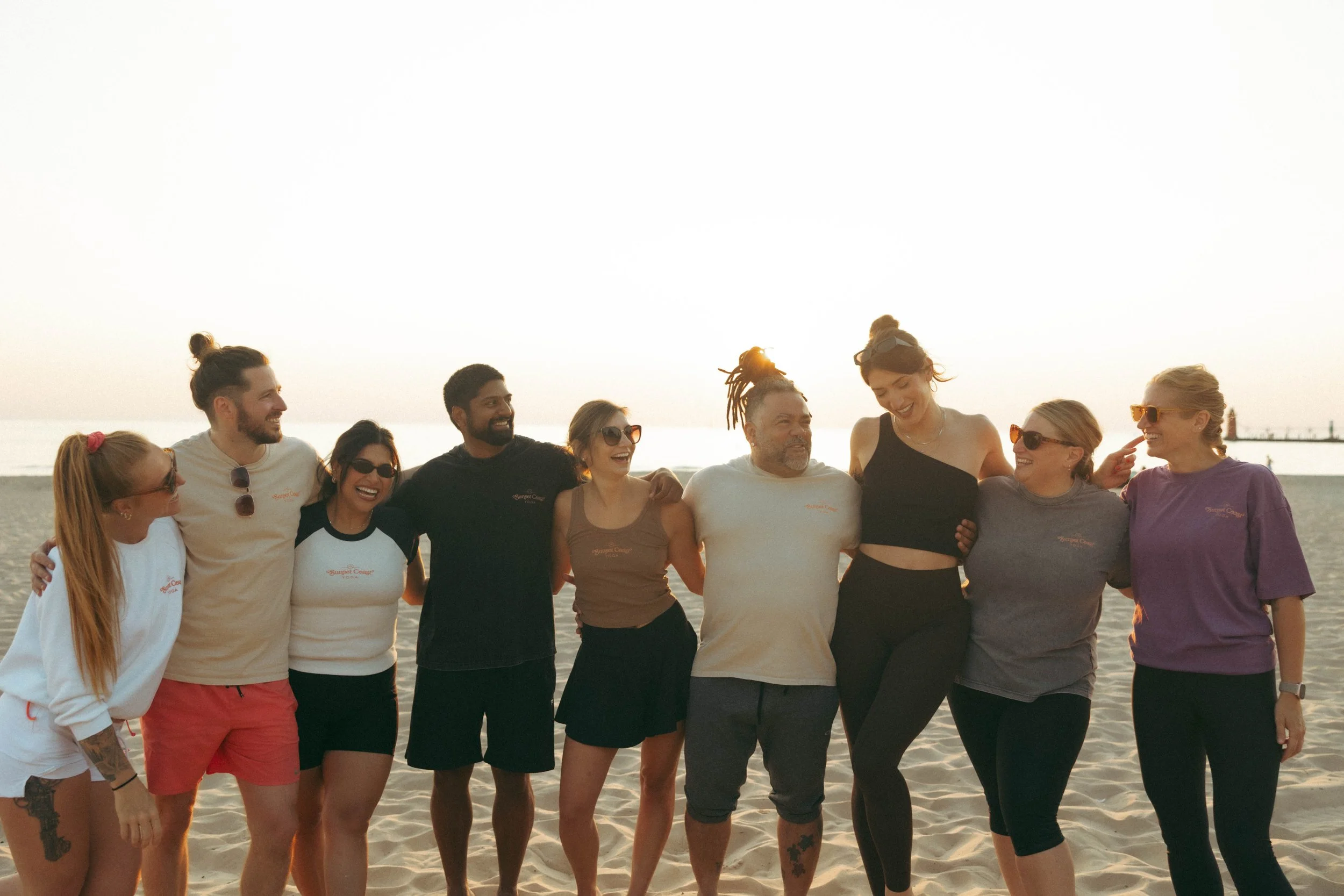 Group of diverse people in casual beachwear standing together on the sand during sunset, smiling and with arms around each other.