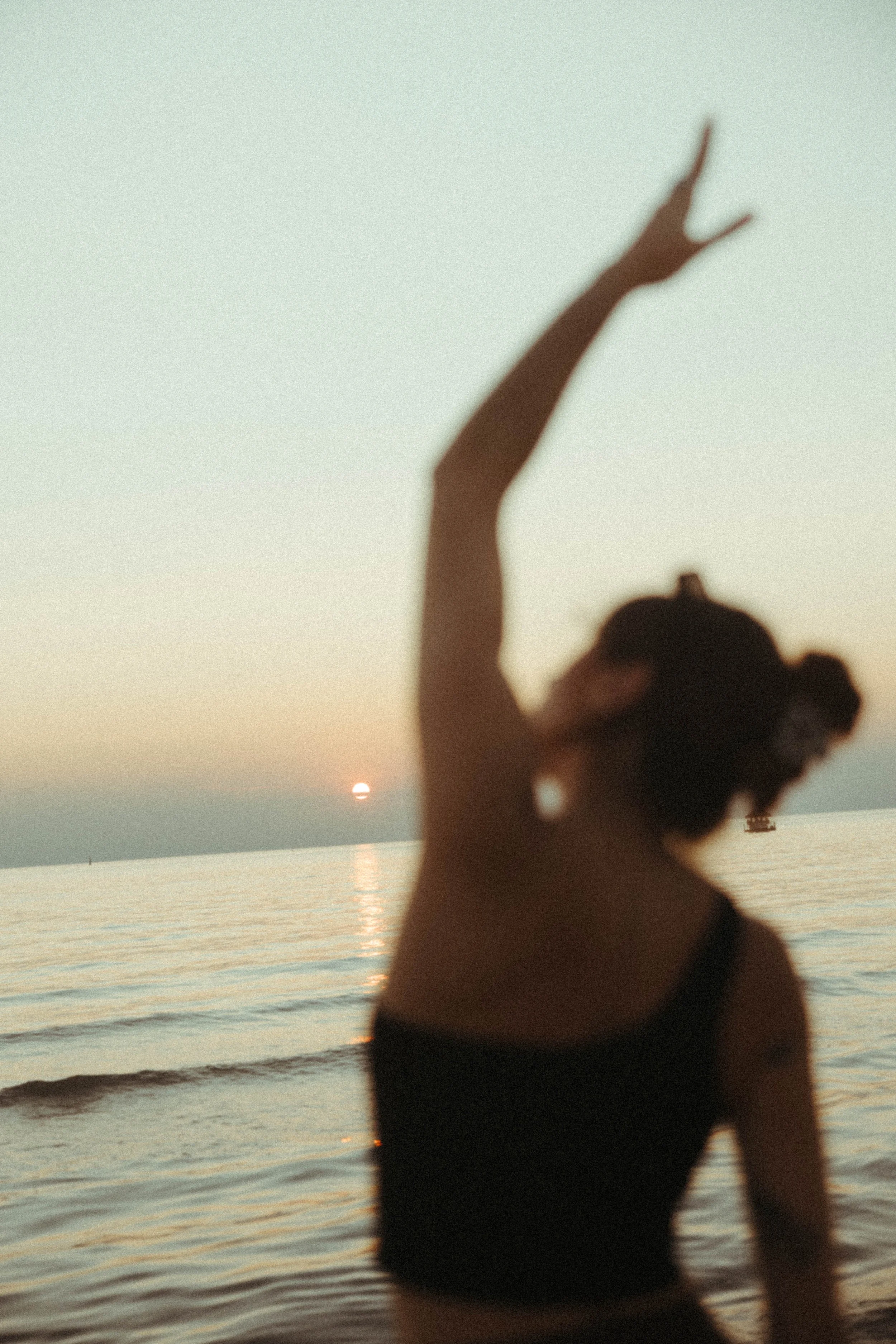 Silhouette of a person with hair in buns standing on a beach with the sun setting over the ocean, casting a reflection on the water.