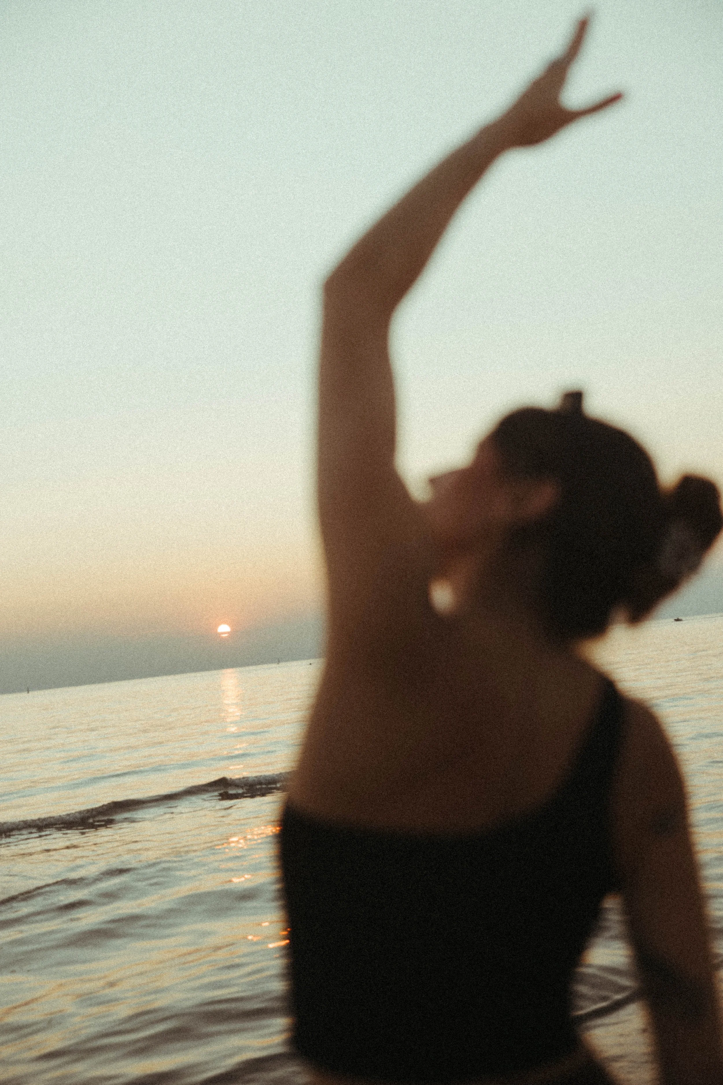 A person standing at the beach during sunset, with their back turned and arm raised high, holding a V-shaped hand gesture. The sky is clear with the sun setting over the horizon, casting reflections on the water.