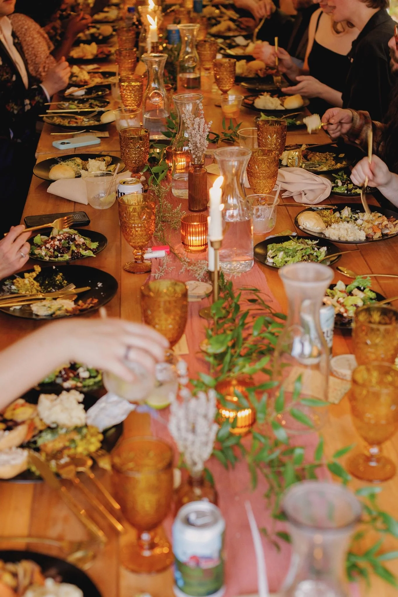 A long wooden dining table with multiple people gathered around, eating a meal. The table is decorated with candles, vases, and greenery. Plates with various foods, drinks, and utensils are visible.