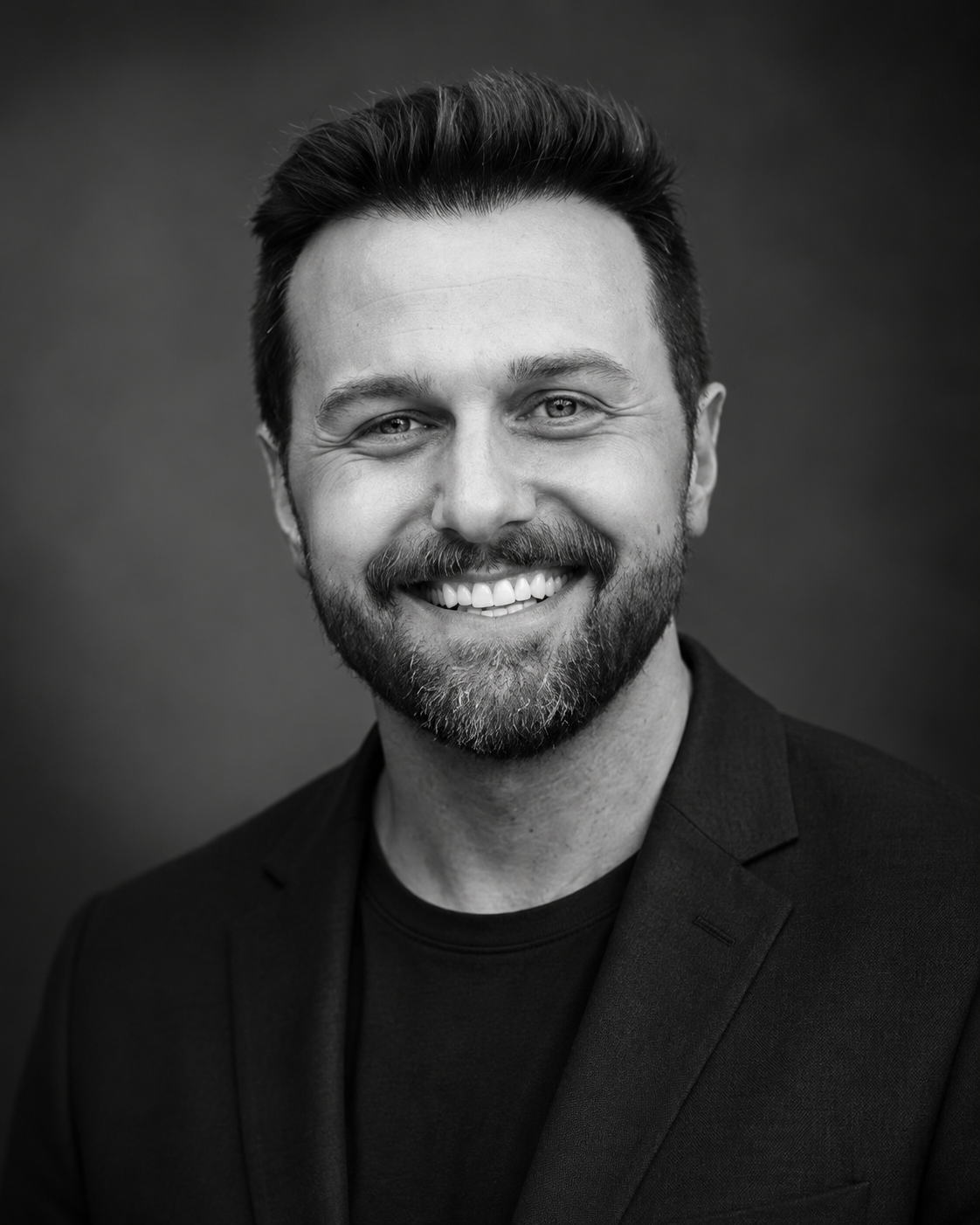 Black and white portrait of a smiling man with a beard, wearing a blazer and a shirt.