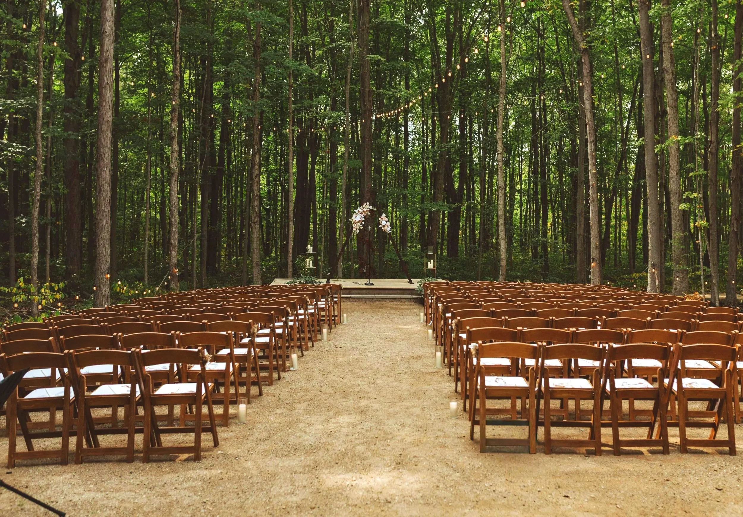 Outdoor wedding ceremony setup in a forest with rows of wooden chairs on either side of a dirt aisle, decorated with candles, and a small stage with floral arrangements and a microphone, surrounded by tall trees with string lights overhead.