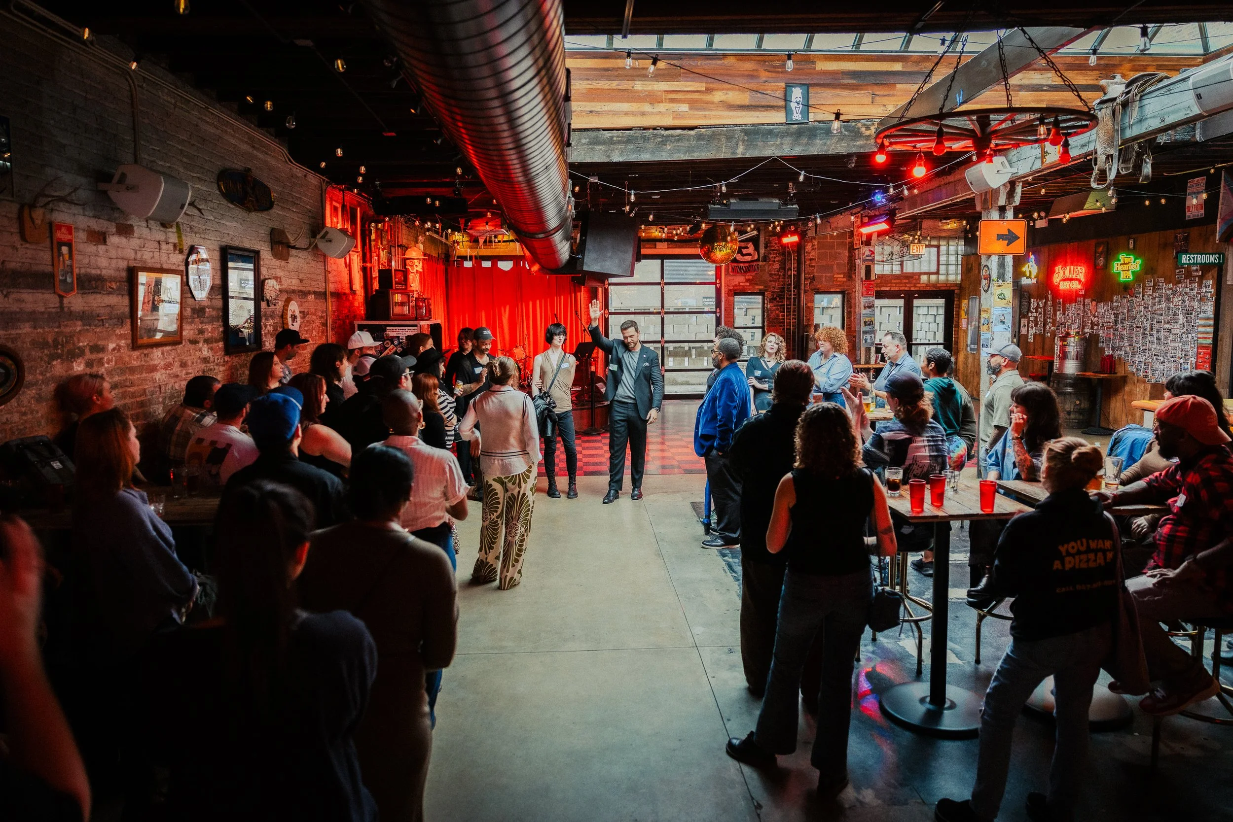 People attending an indoor event with a person speaking or performing on stage, red curtains, and eclectic decor, in a dimly lit space with exposed brick walls.