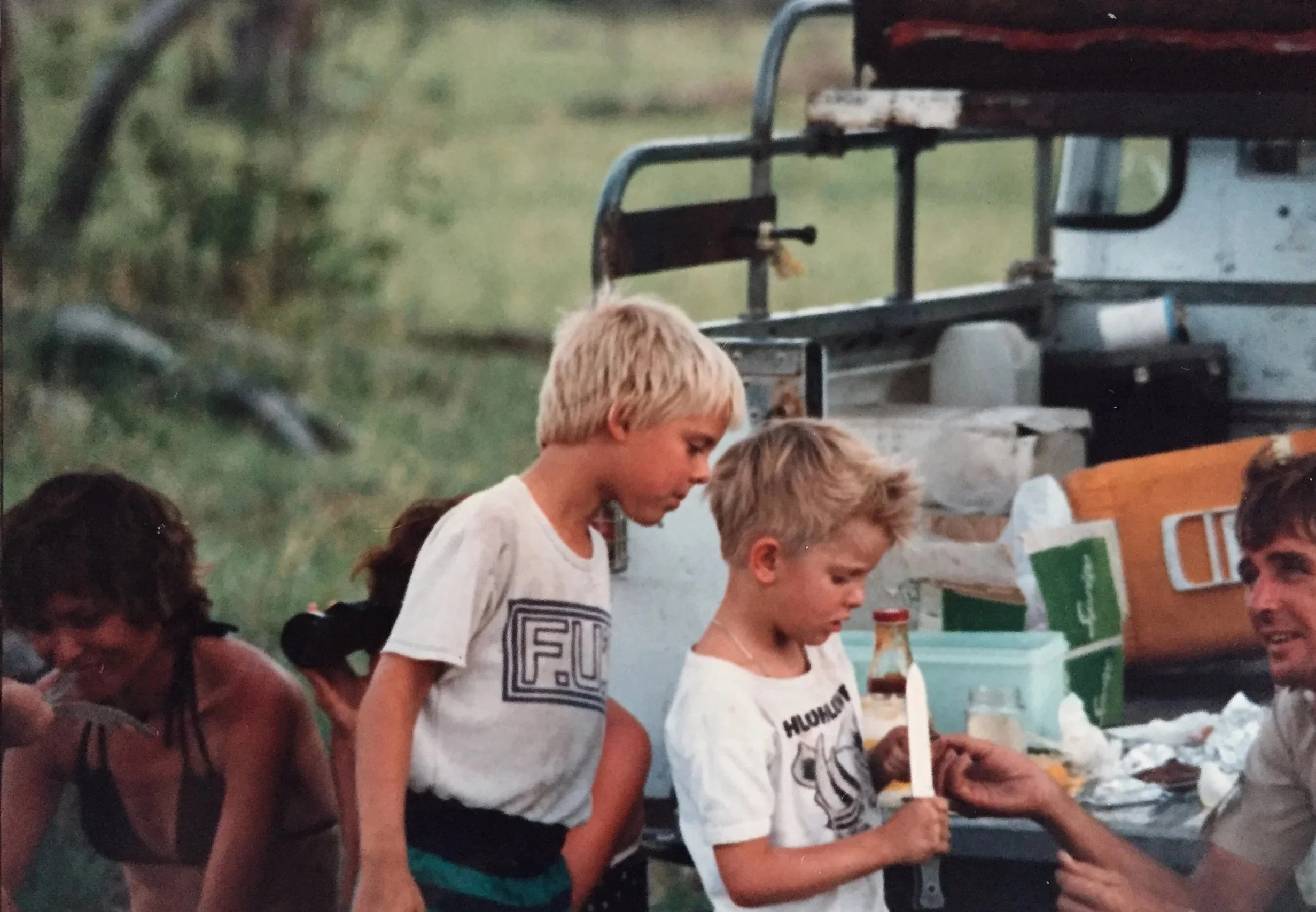 A vintage 1980s safari camp scene in the Okavango Delta, where two young blonde boys stand by the open back of a Land Rover while a man guides one boy holding a safari knife near a tailgate spread of food.