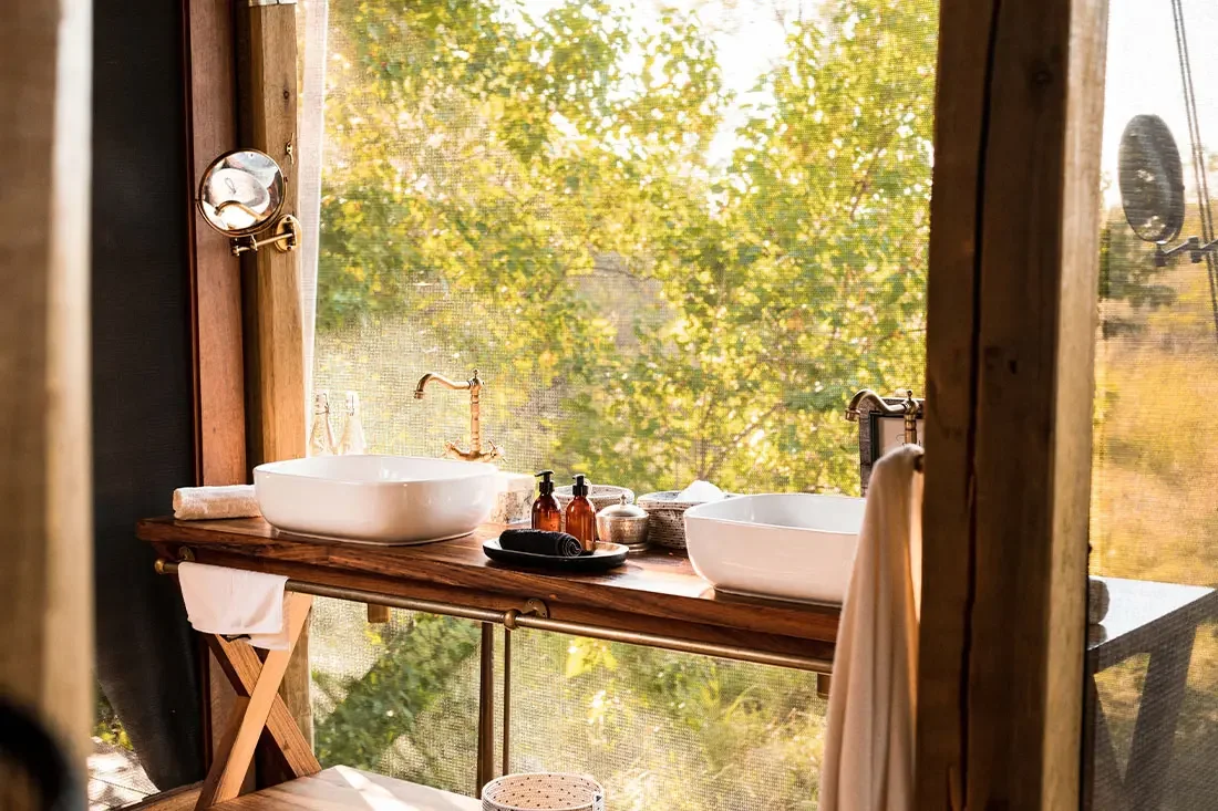 A modern double vanity bathroom with white vessel sinks set against a large glass window overlooking bright green trees.