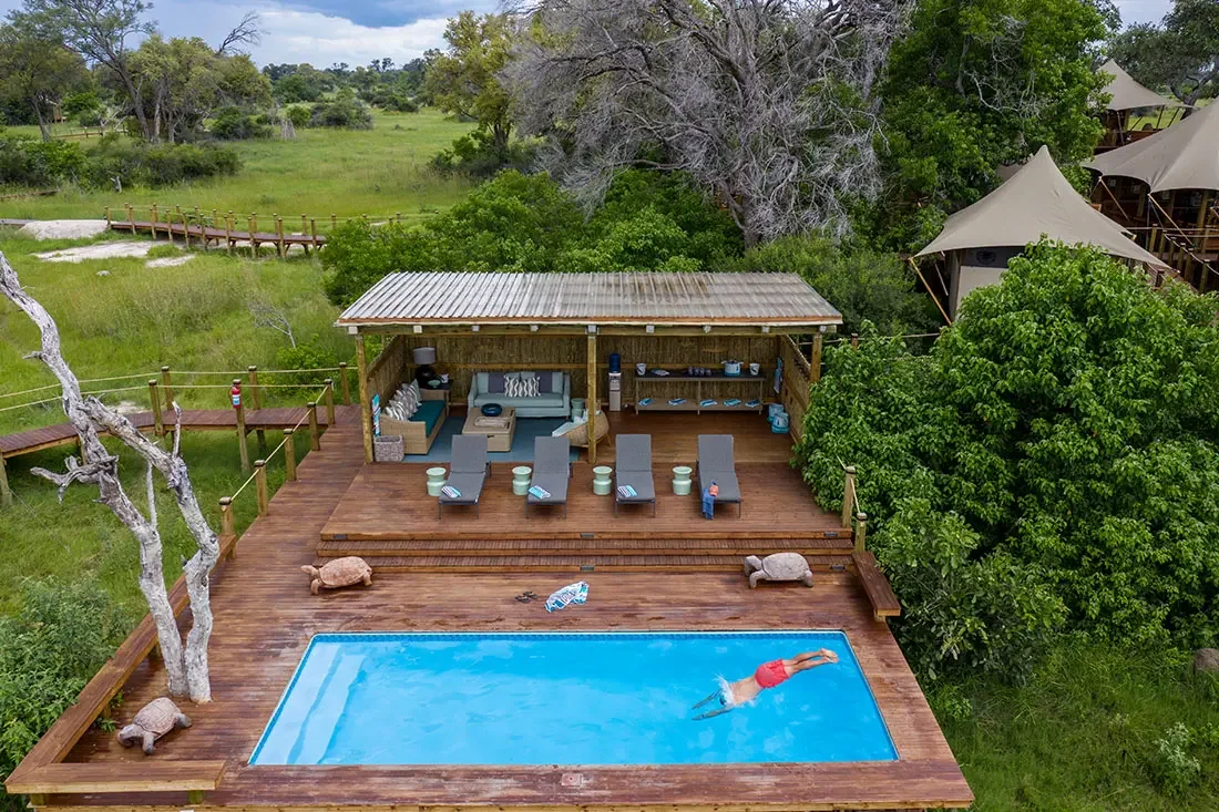 An aerial view of a rectangular blue swimming pool embedded in a wooden deck, complete with lounge chairs, a shaded pool house, and surrounding trees.