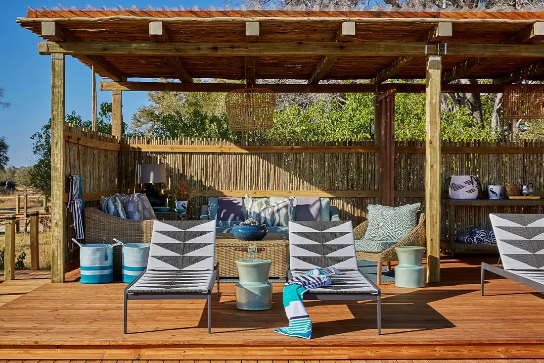 A sunny wooden deck lounge protected by a reed wall, featuring striped lounge chairs, a woven couch with cushions, and blue decorative barrels.