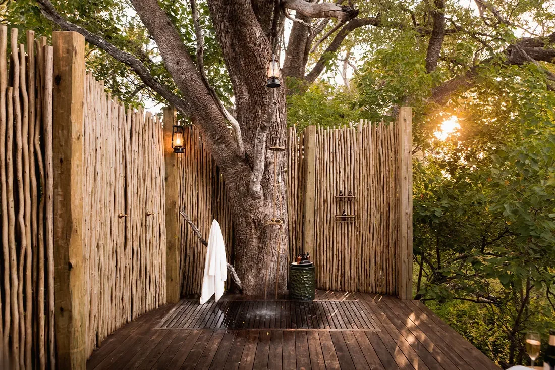 An outdoor shower built against the trunk of a large tree, enclosed by high rustic wooden stick walls.