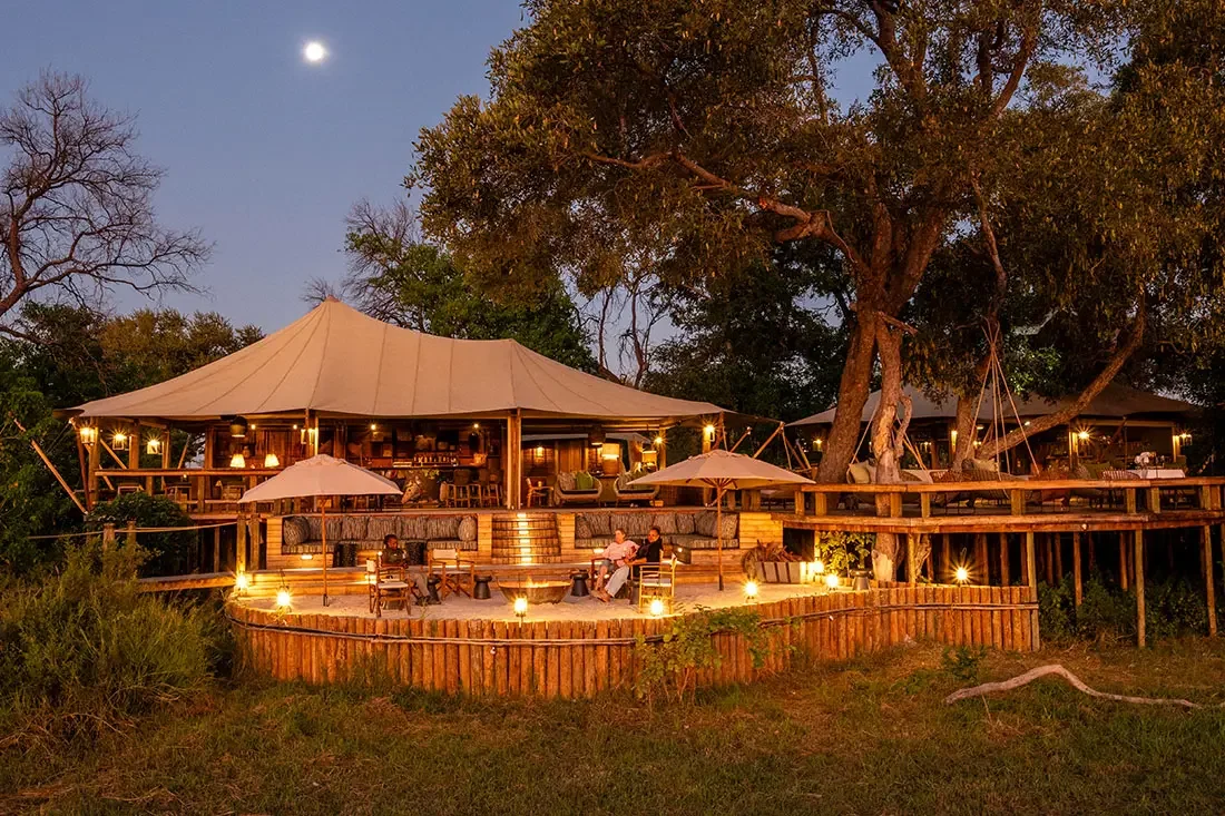 A large tented safari lodge illuminated at night, featuring a circular wooden deck with outdoor seating under a moonlit sky.
