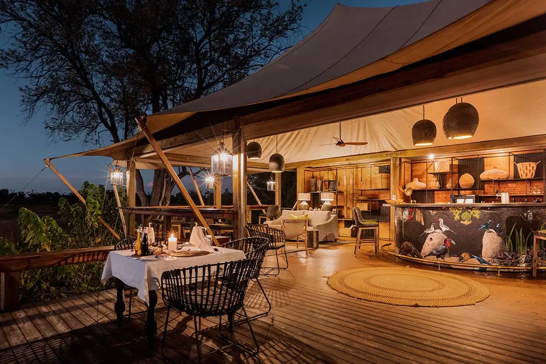 An illuminated outdoor dining deck at night featuring a set dinner table, lounge couches, and a decorated circular bar under a canvas canopy.