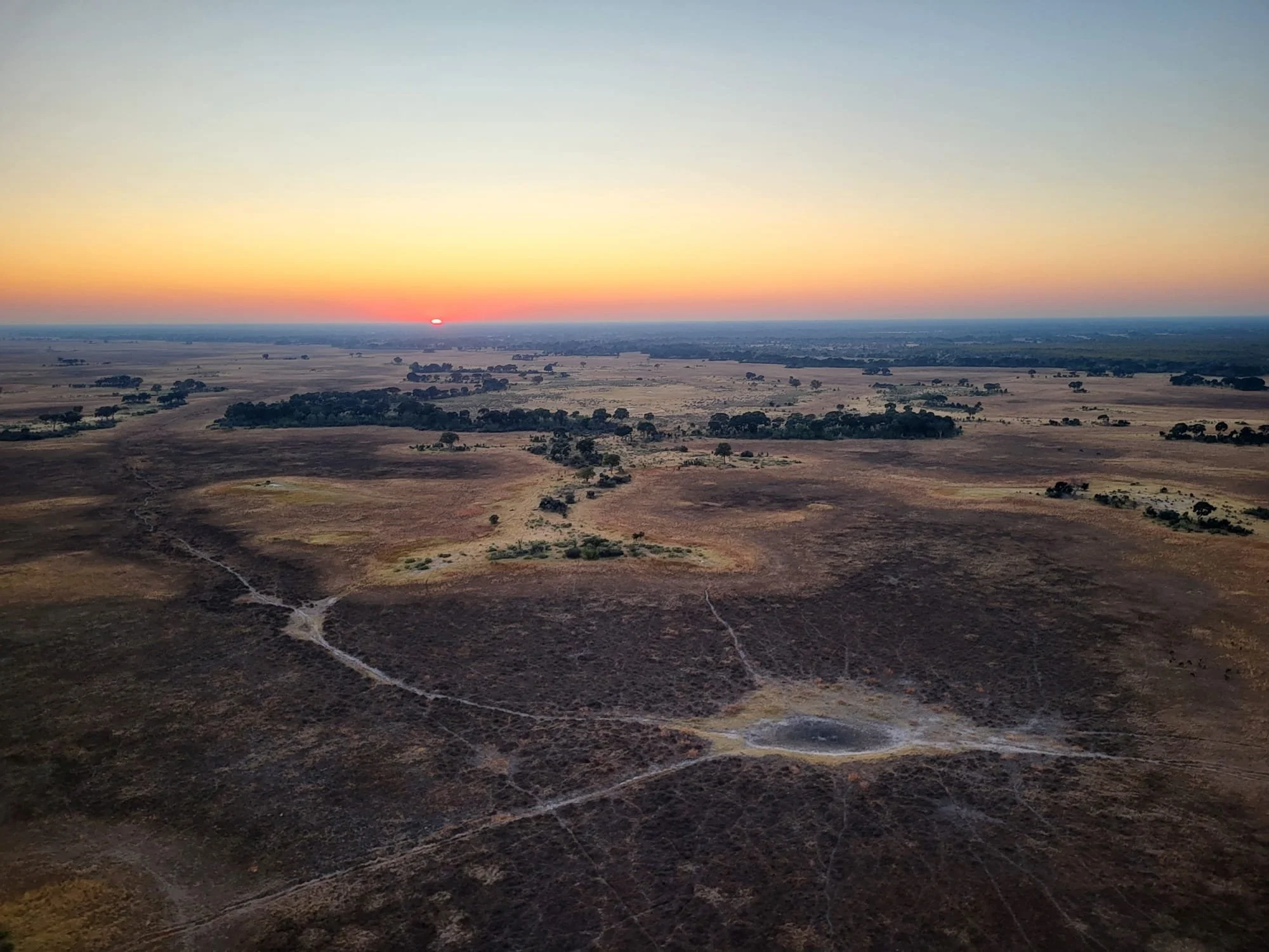 An aerial view of a vast, flat landscape in the Okavango Delta at sunrise with a network of dry paths snaking across the ground.