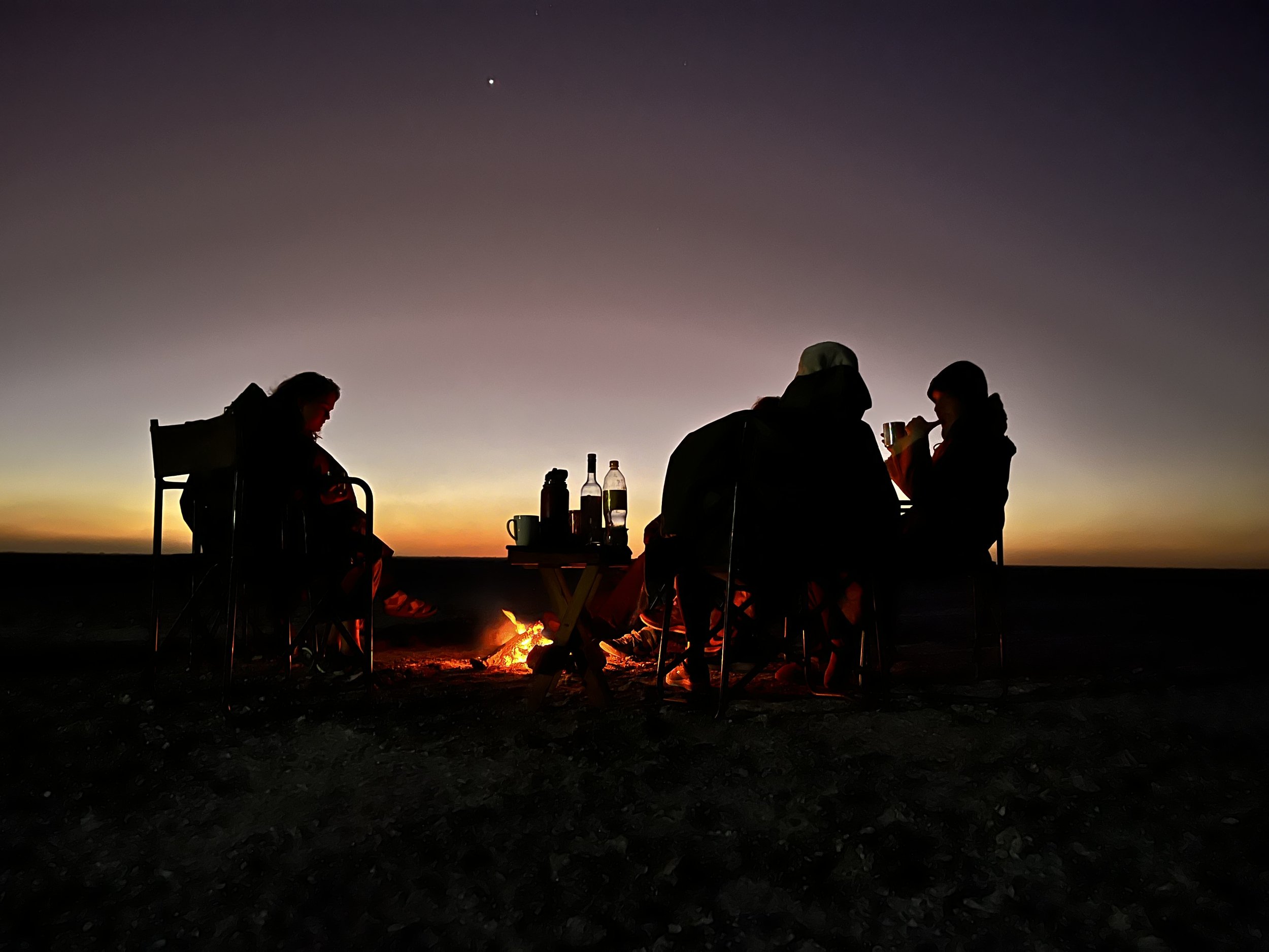 Three people sitting around a fire as the last rays of sunshine disappear in the Makgadikgadi Salt Pans, Botswana.