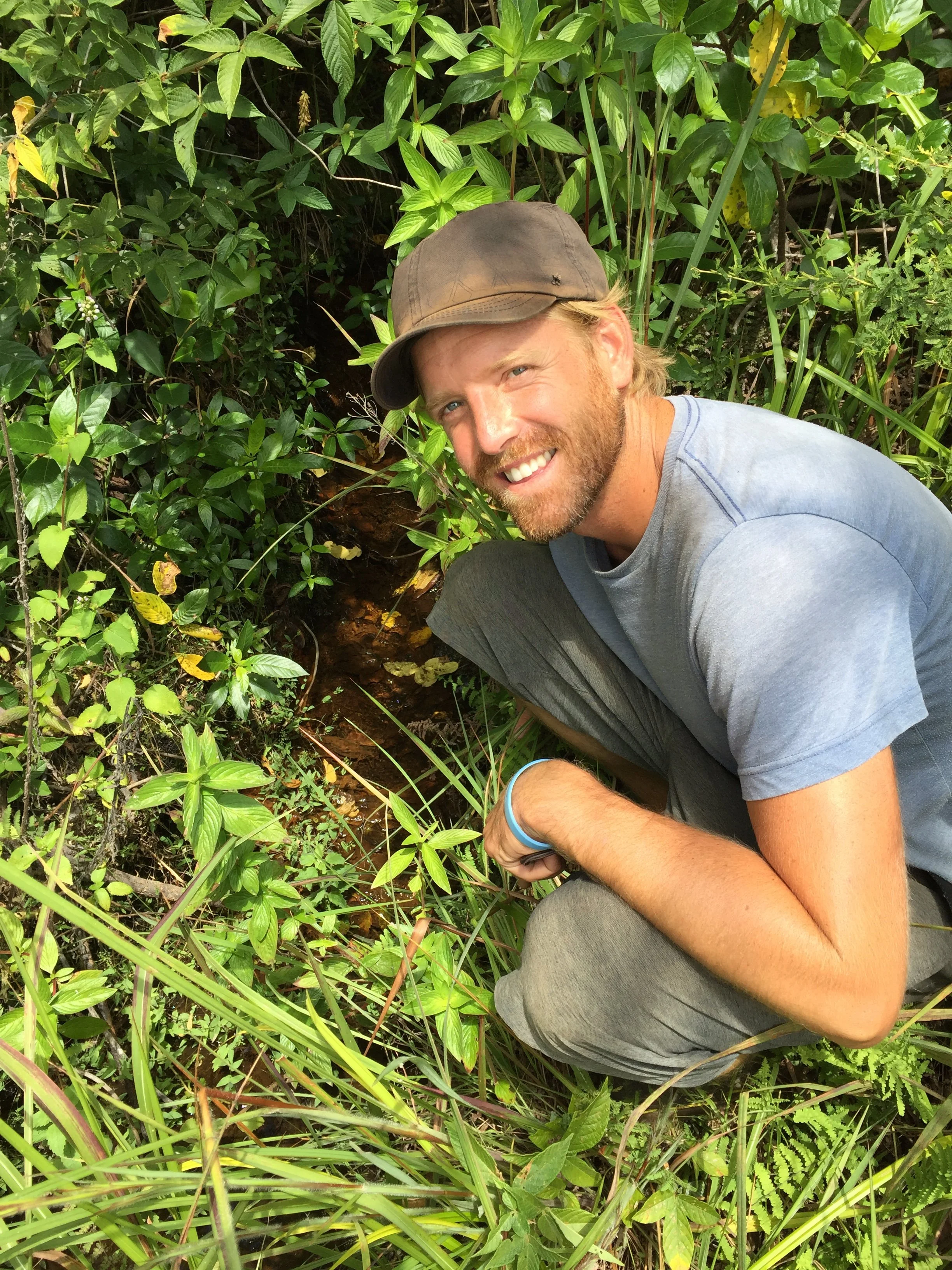 A smiling man wearing a baseball cap crouches among dense green leaves and grass at the natural source of the Okavango River in Angola.