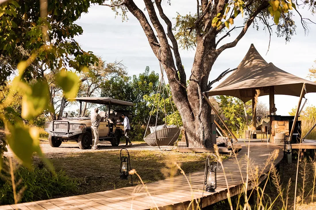 A game drive vehicle parked on a dirt path next to a raised wooden walkway and an open-air safari tent under large trees.