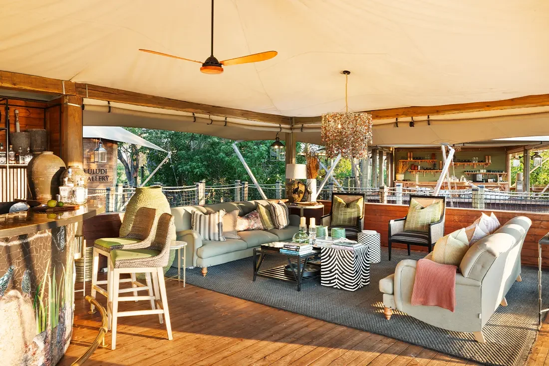 Luxurious safari lodge interior with plush white couches, patterned pillows, a bar area, and open walls overlooking a green landscape.