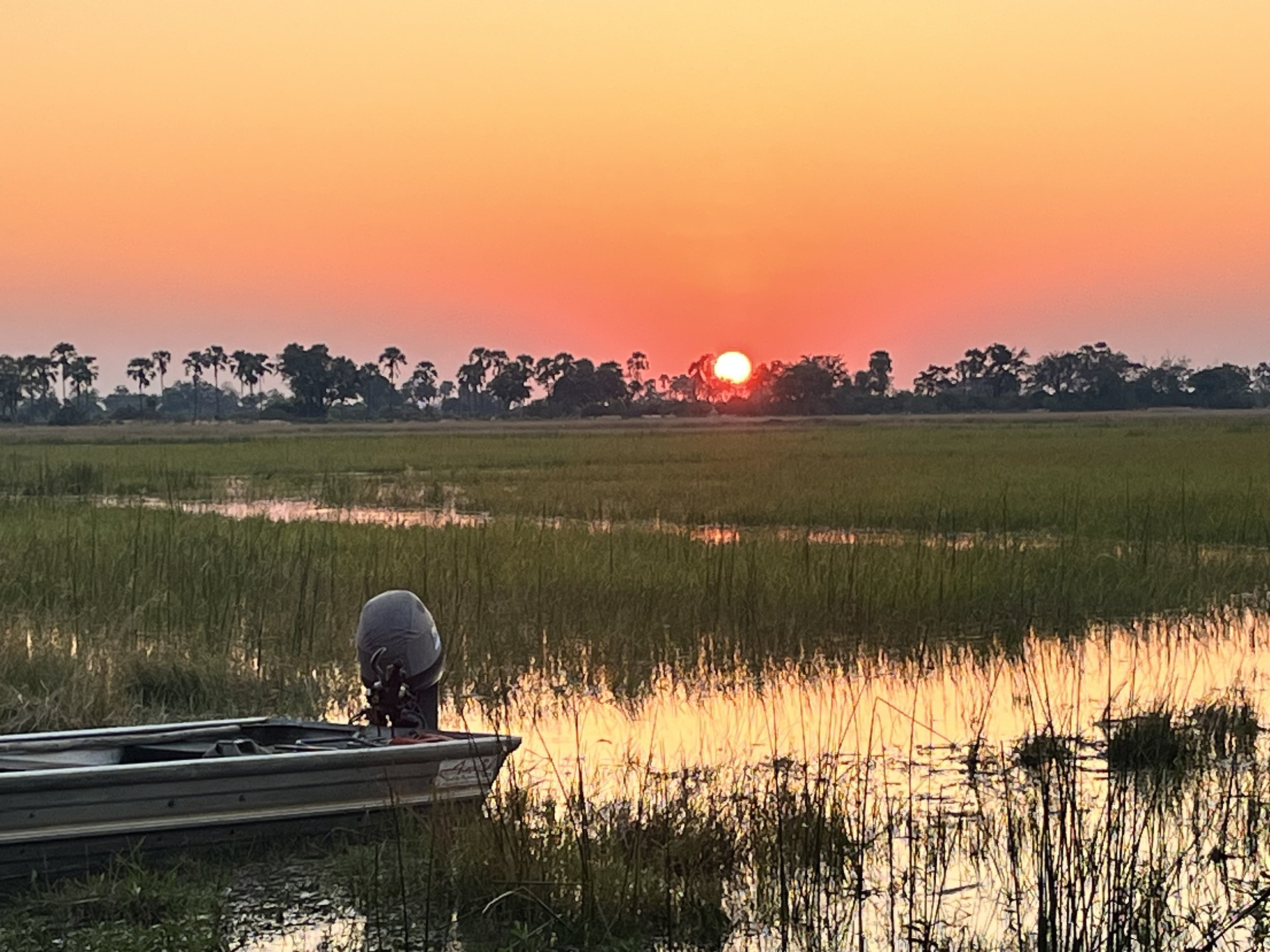 A small safari boat sits in a quiet river channel as the sun sets over a wide floodplain in the Okavango Delta.