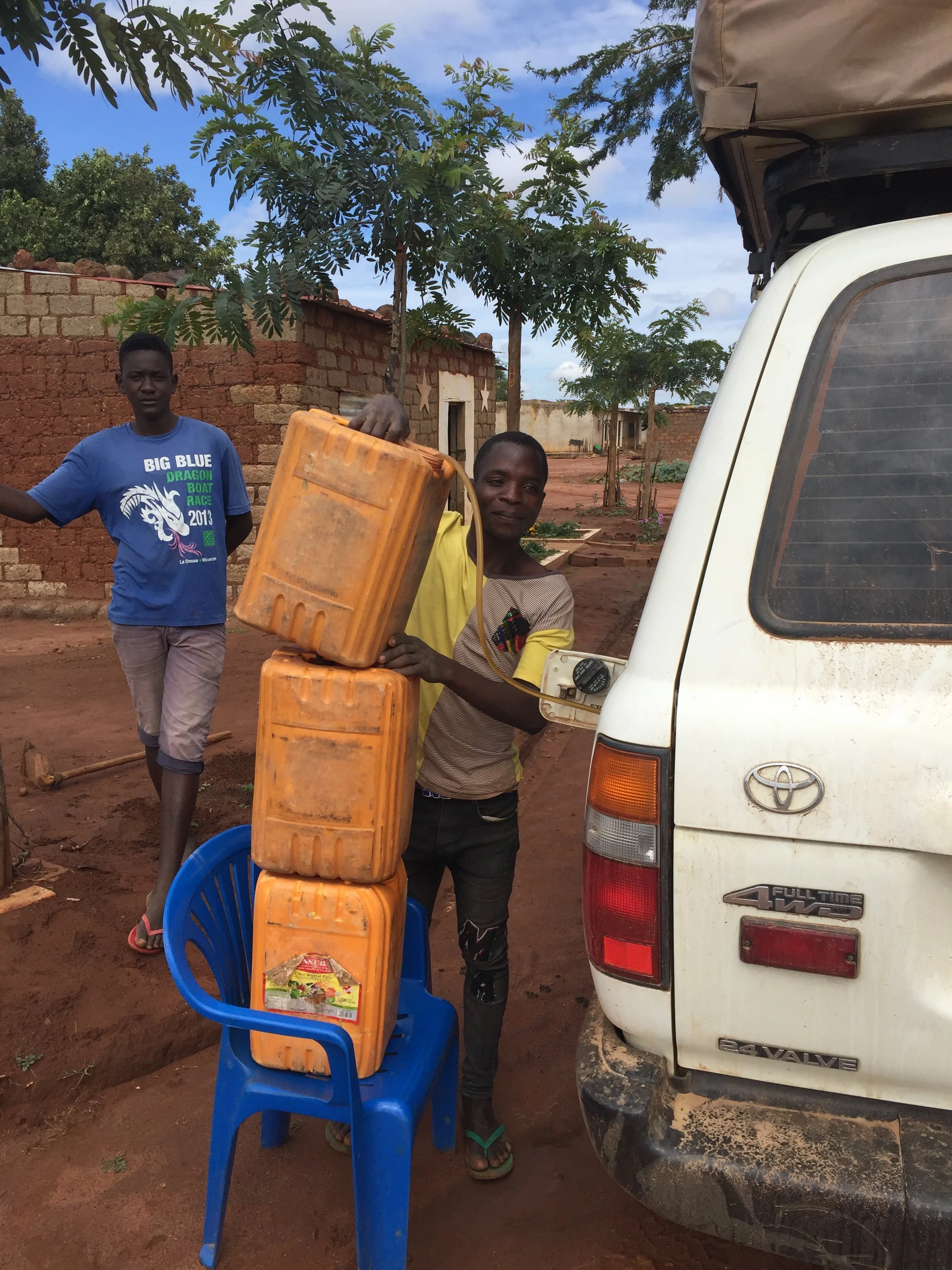 Two local men stack orange plastic jerrycans on a blue chair next to a white Toyota Land Cruiser during an overland trip in Angola.