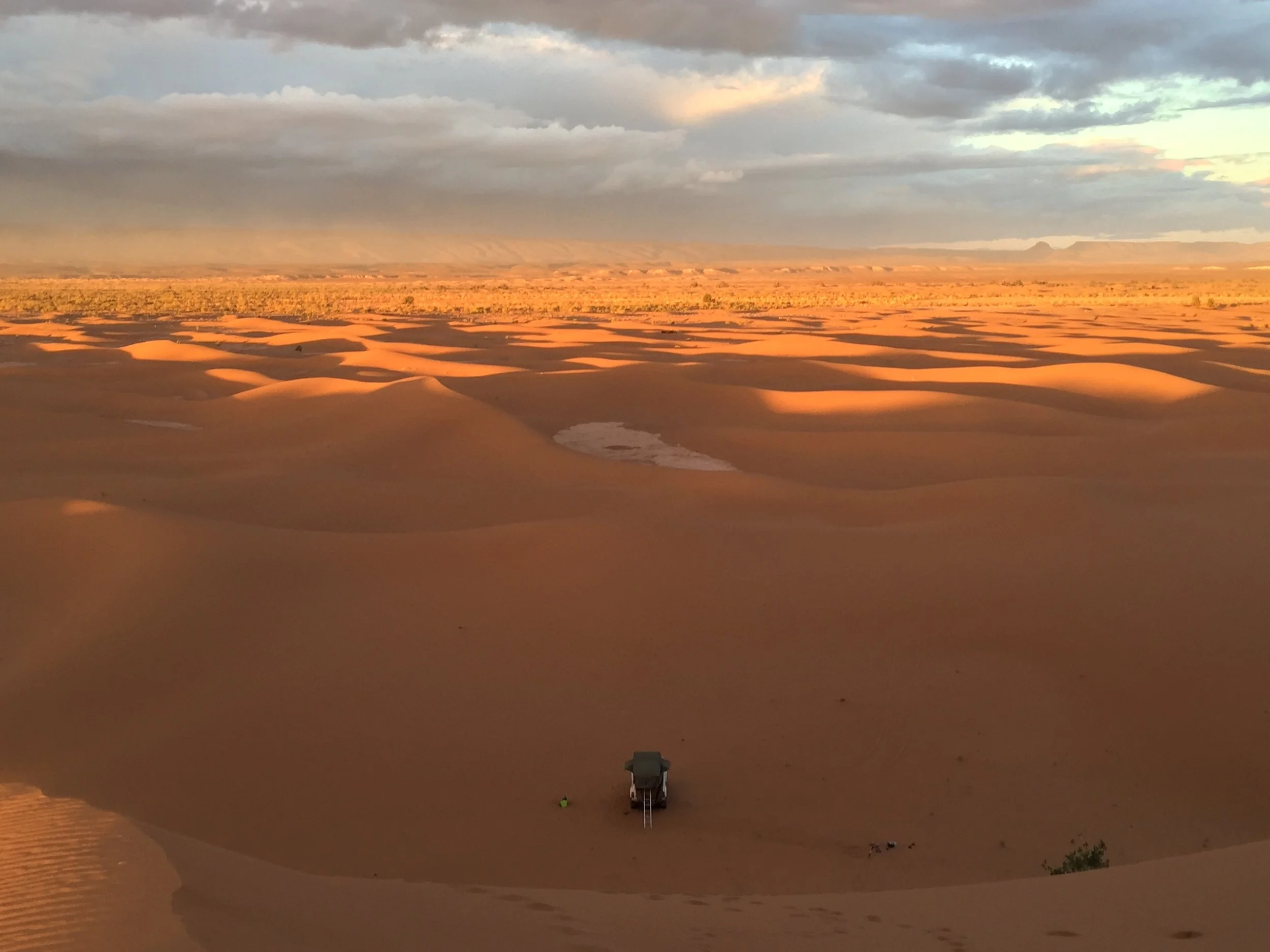 A high-angle scenic view looking across rolling orange sand dunes toward the Algerian border under a dramatic, cloudy sunset sky, with a lone safari vehicle parked at the base of a dune.