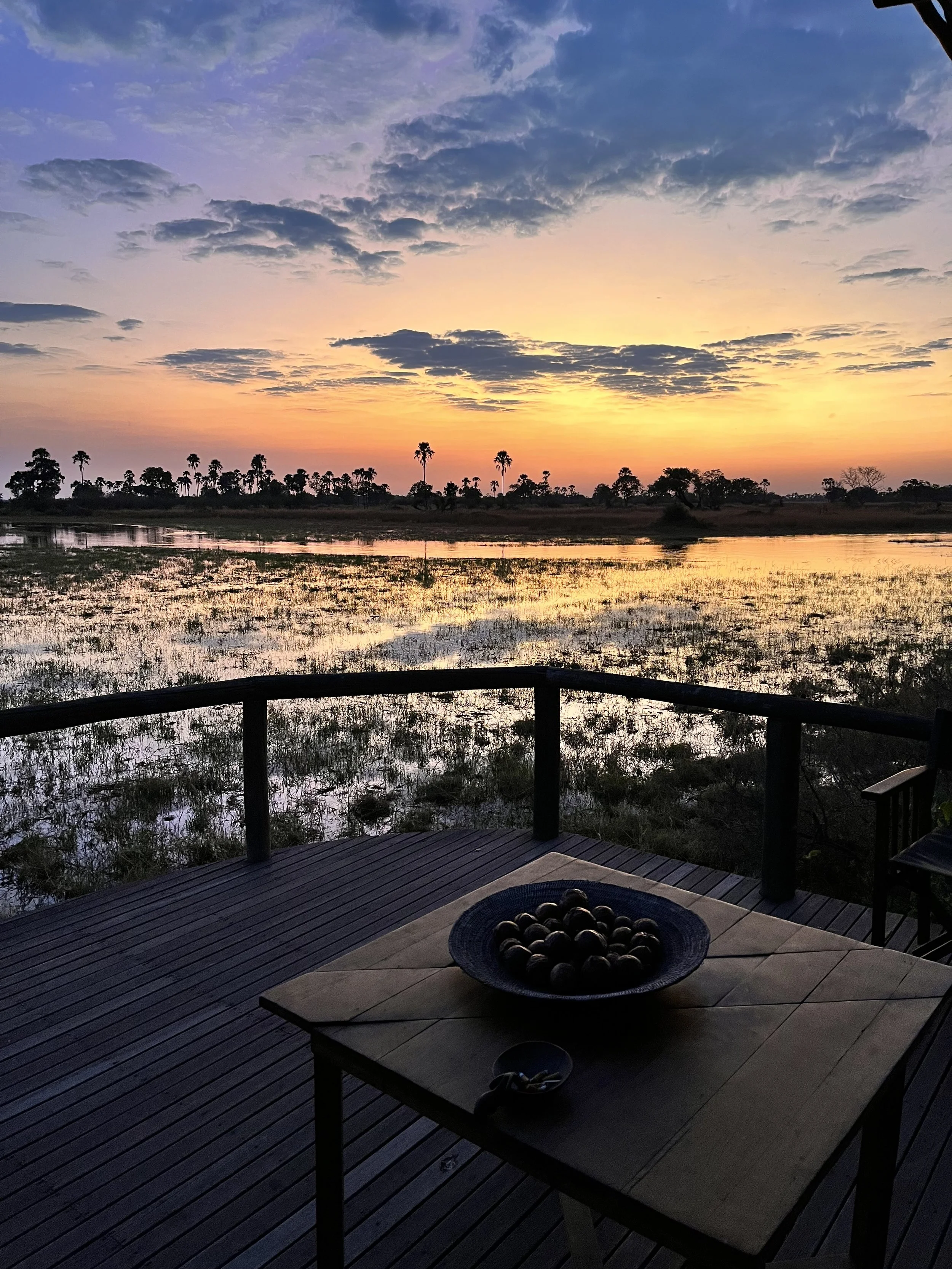 Viewed from an outdoor wooden deck, silhouetted palm trees frame a dark river channel at dusk under a sky filled with oranges and grey-blue colors in the Okavango Delta.