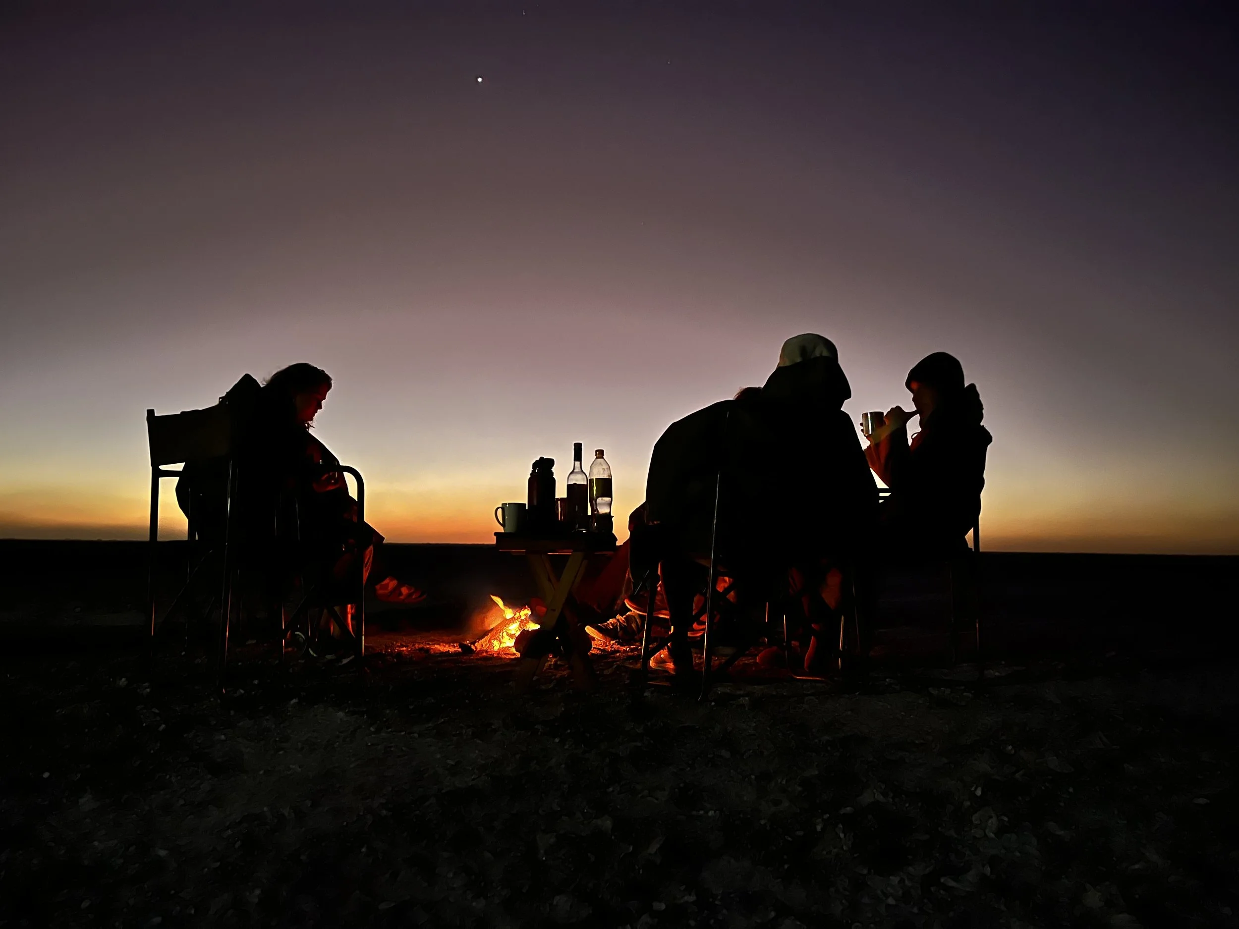 Three people sitting around a fire as the last rays of sunshine disappear in the Makgadikgadi Salt Pans, Botswana