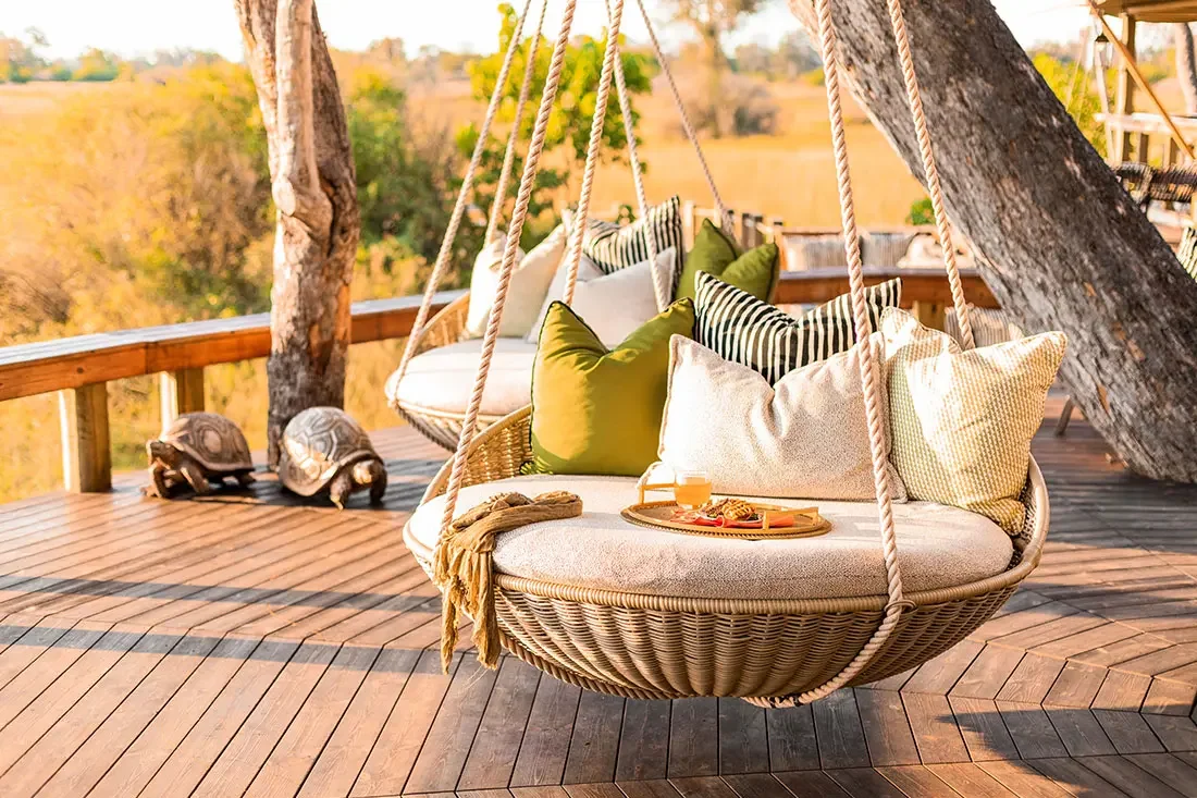 A large, hanging wicker daybed with white cushions and green striped pillows on a wooden deck shaded by trees.