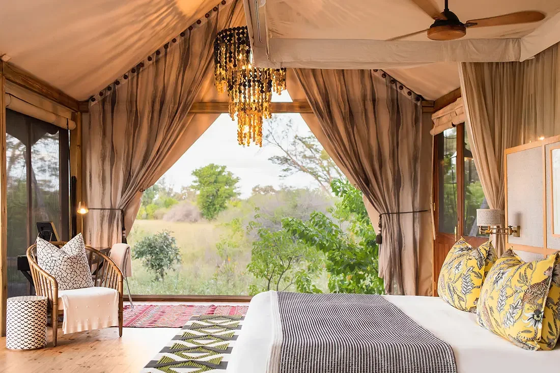 Interior of a tented bedroom looking out through large mesh screen walls toward a sunny, tree-filled landscape.