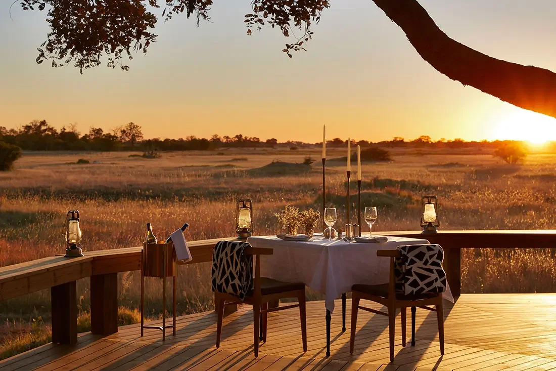 An outdoor dining table set for two on a wooden deck at sunset, overlooking an expansive grassy marshland.