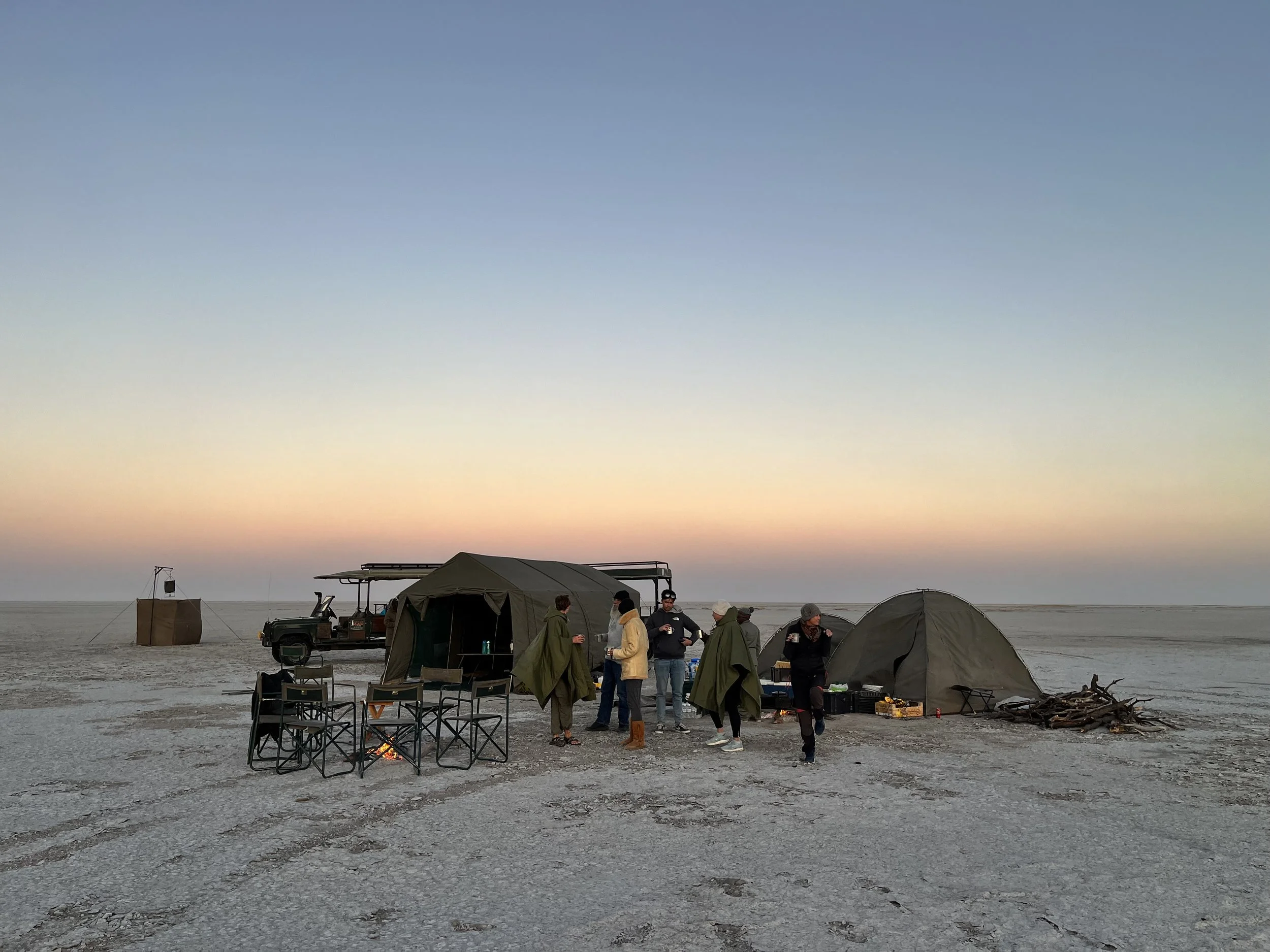 Guests and guides standing around a small mobile safari camp on the Makgadikgadi Salt Pans in Botswana, drinking coffee early in the morning