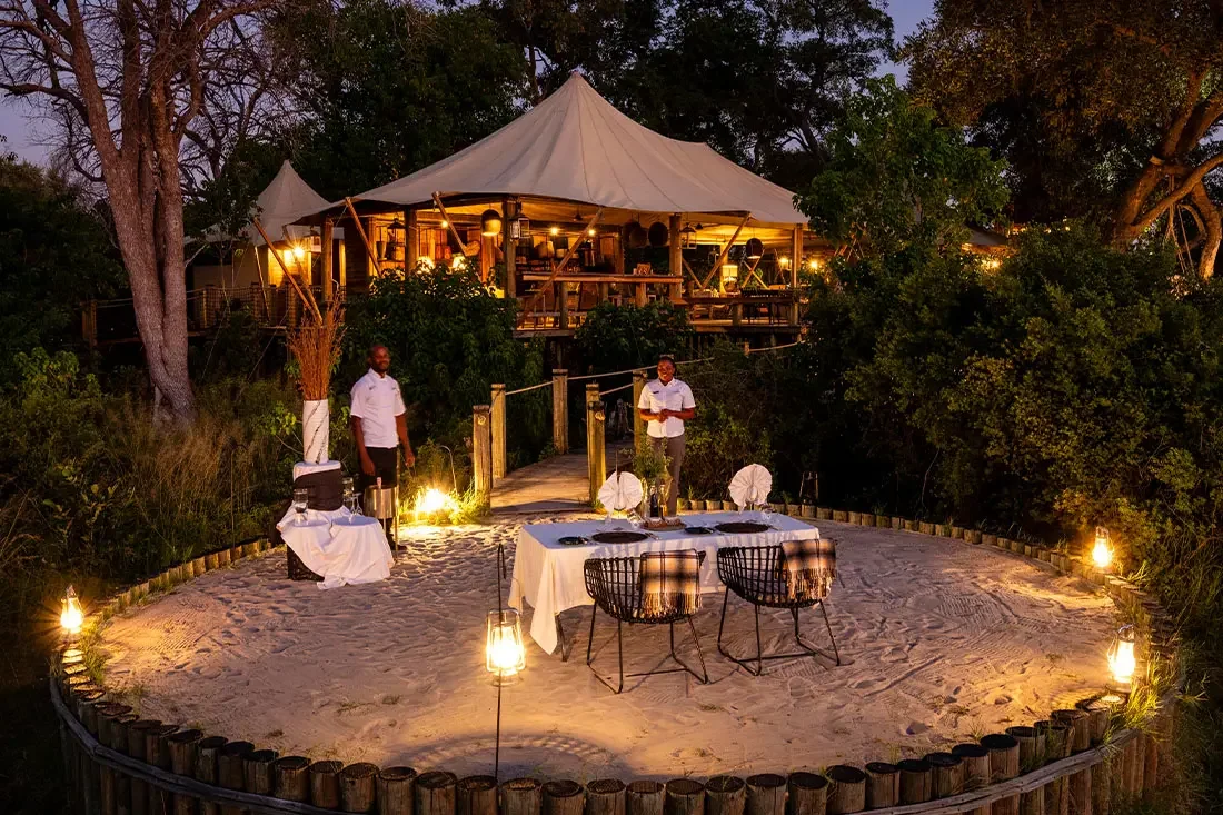 A private outdoor dinner table set for two on a circular sand clearing under a safari tent, with two staff members waiting nearby at dusk.
