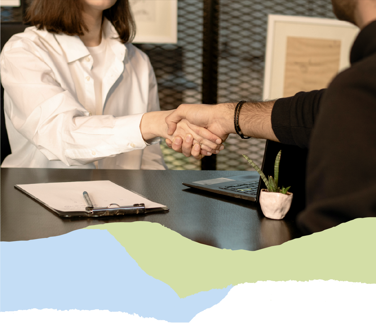 People shaking hands across a table in an indoor setting, with a clipboard and a small potted plant on the table.