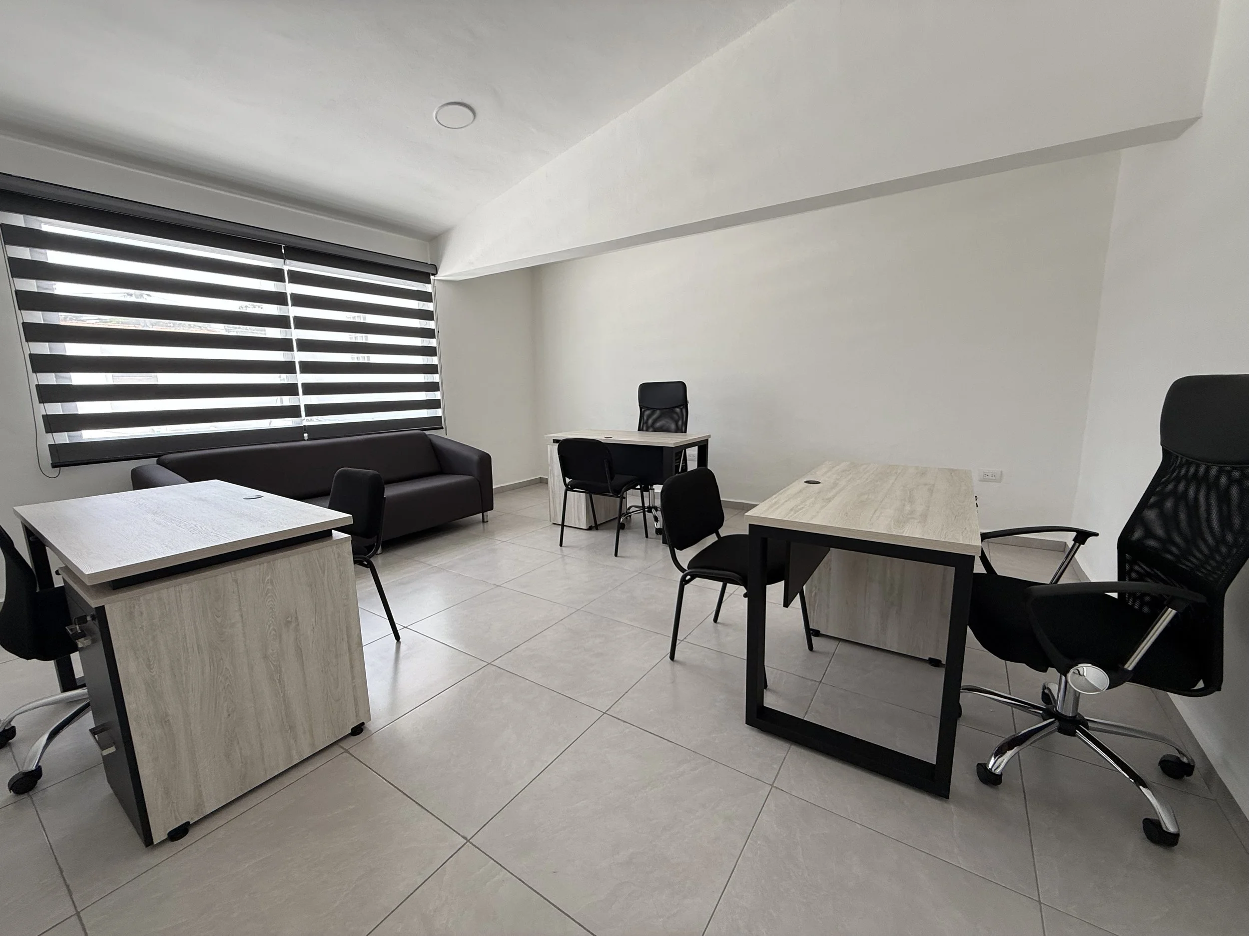 Oficina moderna con escritorios, sillas y un sofá, ventanas con persianas de franjas blancas y negras, paredes blancas y suelo de azulejos.