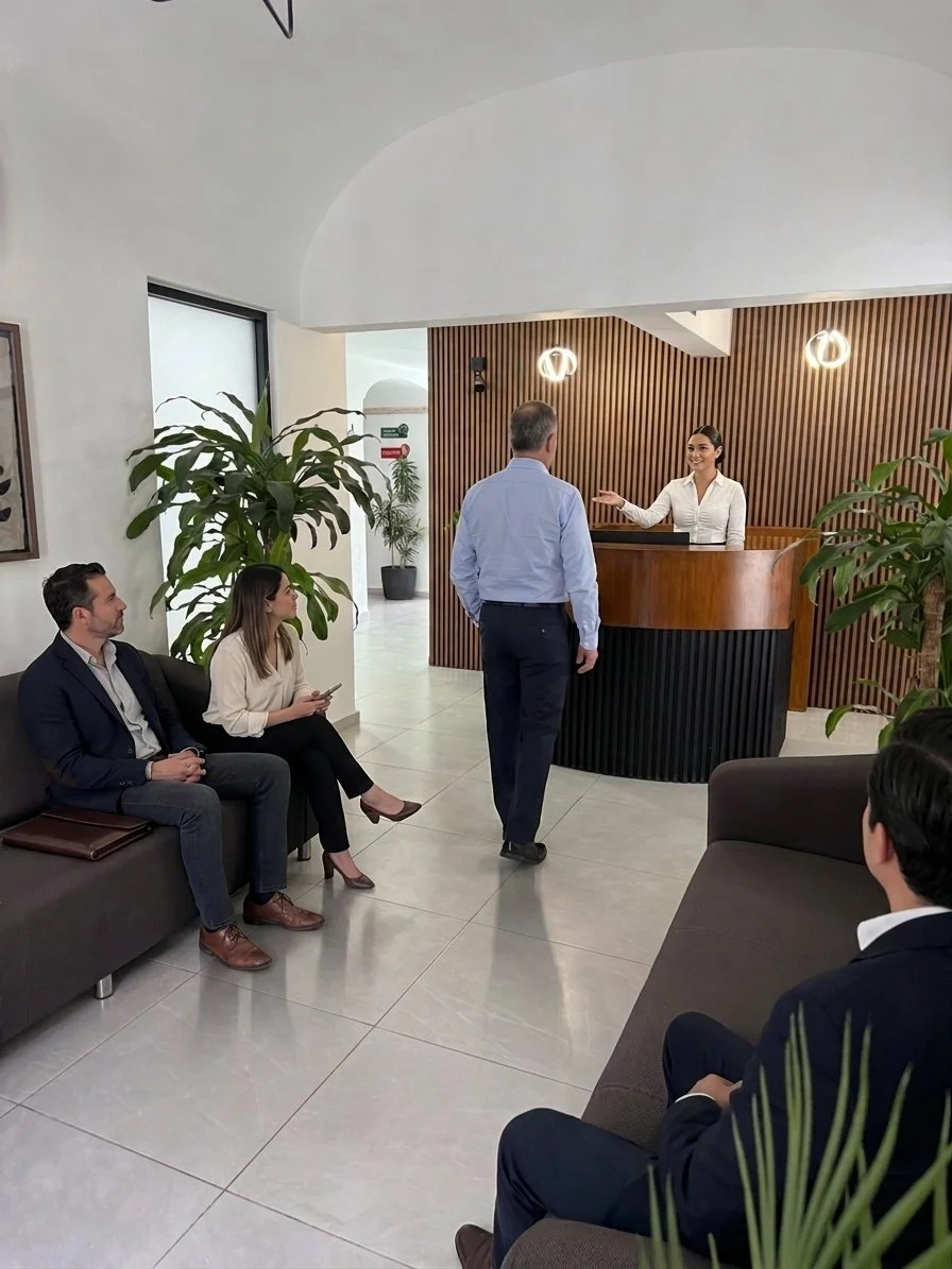 Recepcionista atendiendo a un cliente en la recepción de una oficina, con otras tres personas sentadas esperando ser atendidas